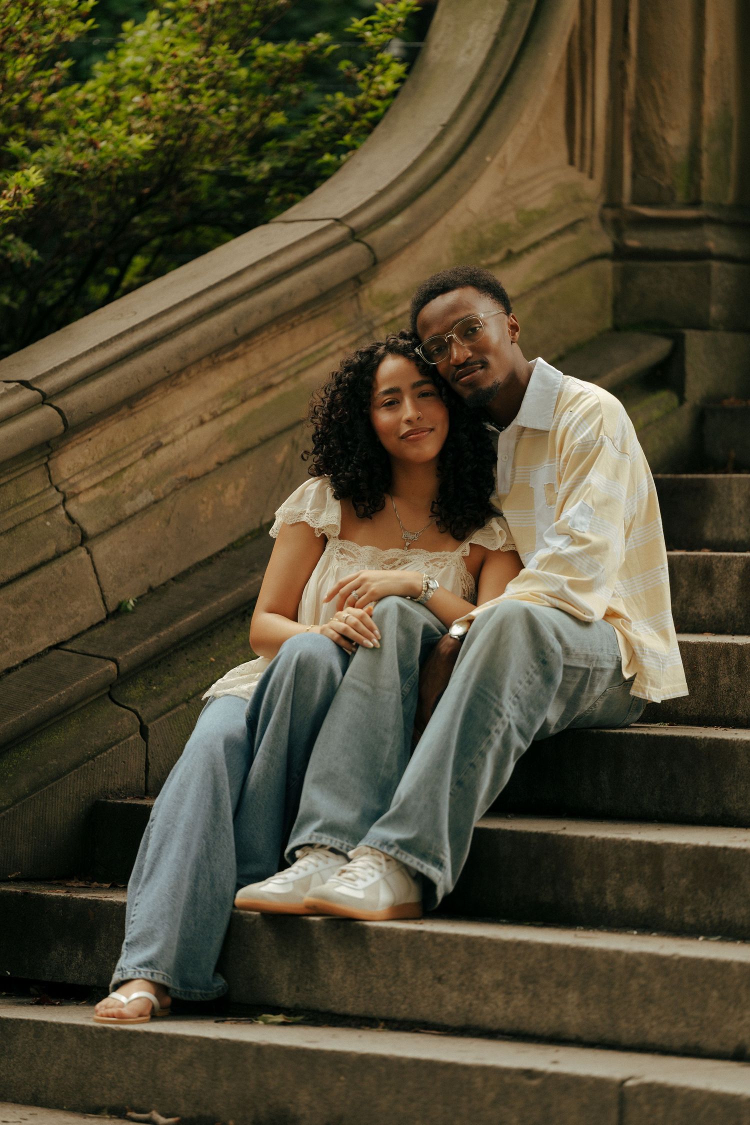 A couple sits closely together on stone steps, smiling at the camera, surrounded by lush greenery.