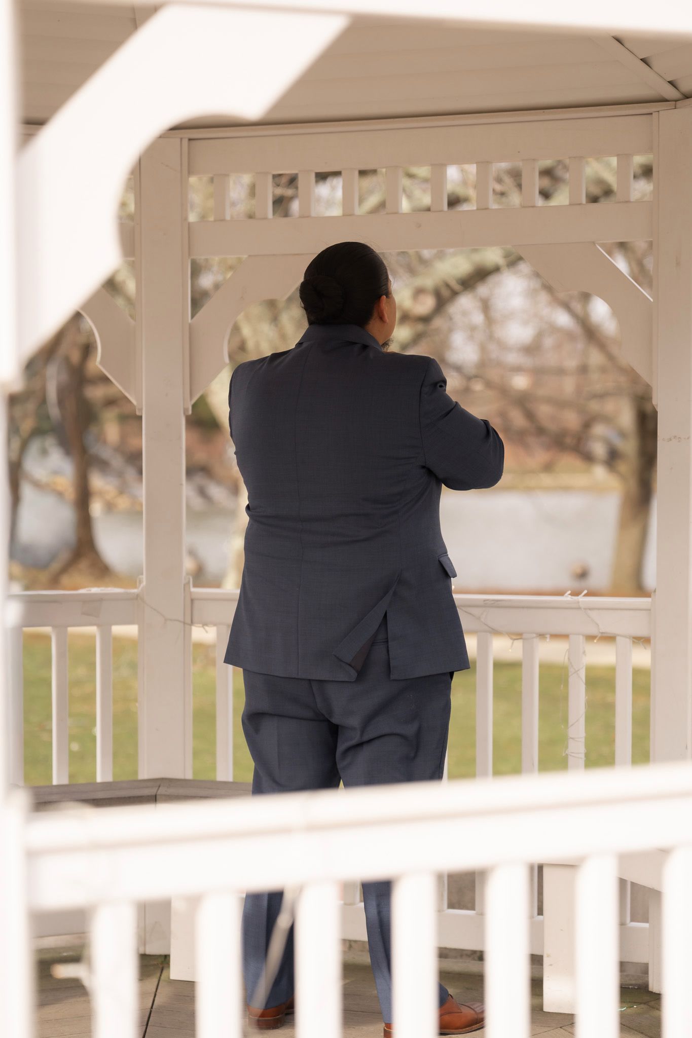 Man in suit standing inside white gazebo, viewed from behind, looking out at park trees.