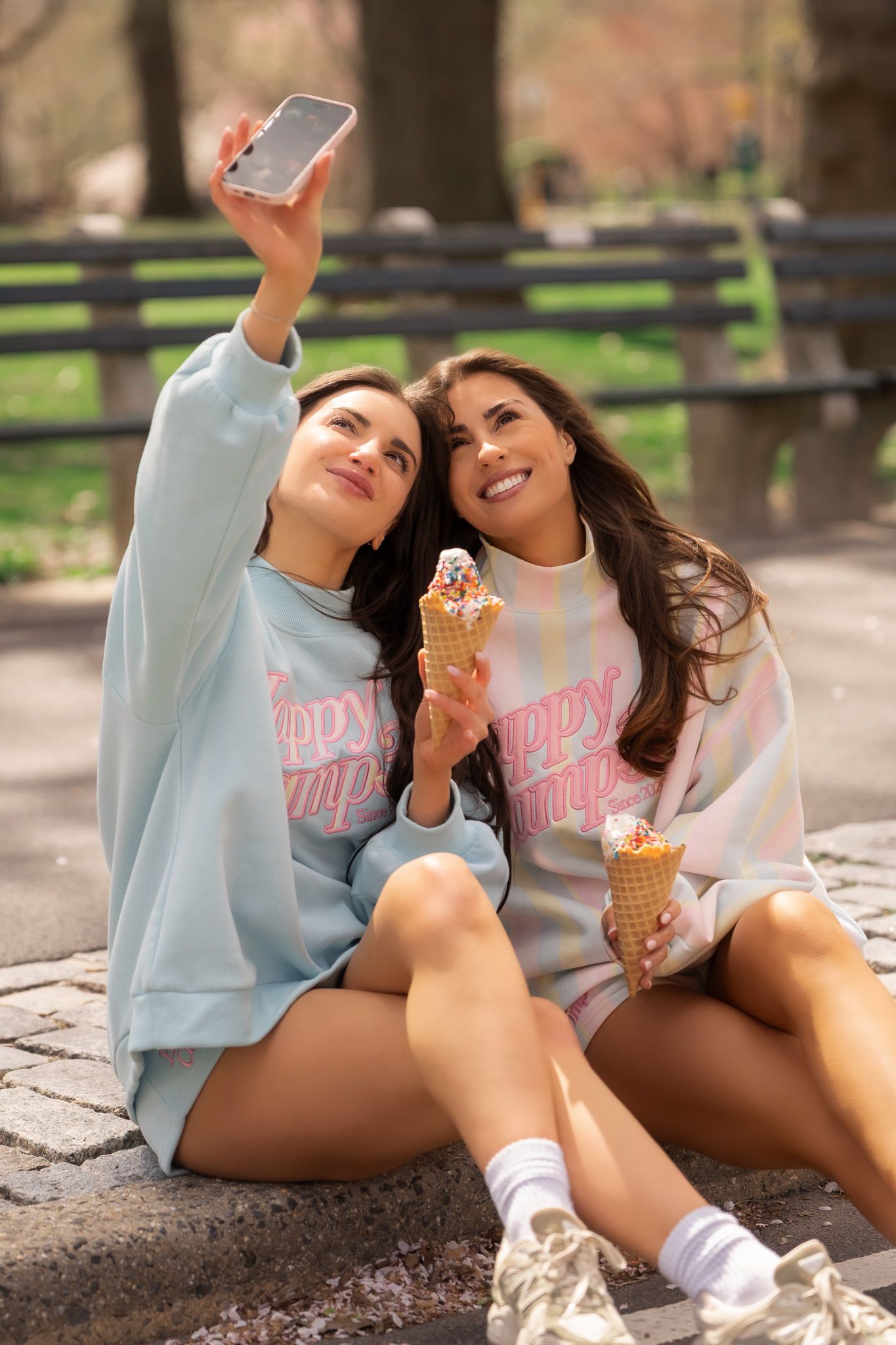 Two young women in matching pastel sweatshirts take a selfie while enjoying ice cream cones in a sunny park.