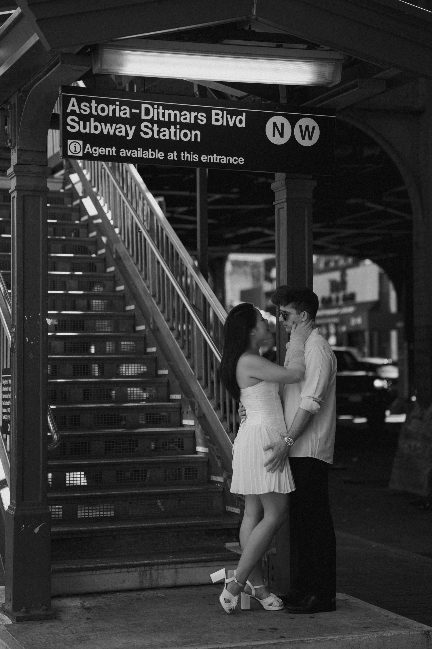 Couple sharing a romantic kiss at Astoria-Ditmars Blvd subway station entrance in black and white photo.