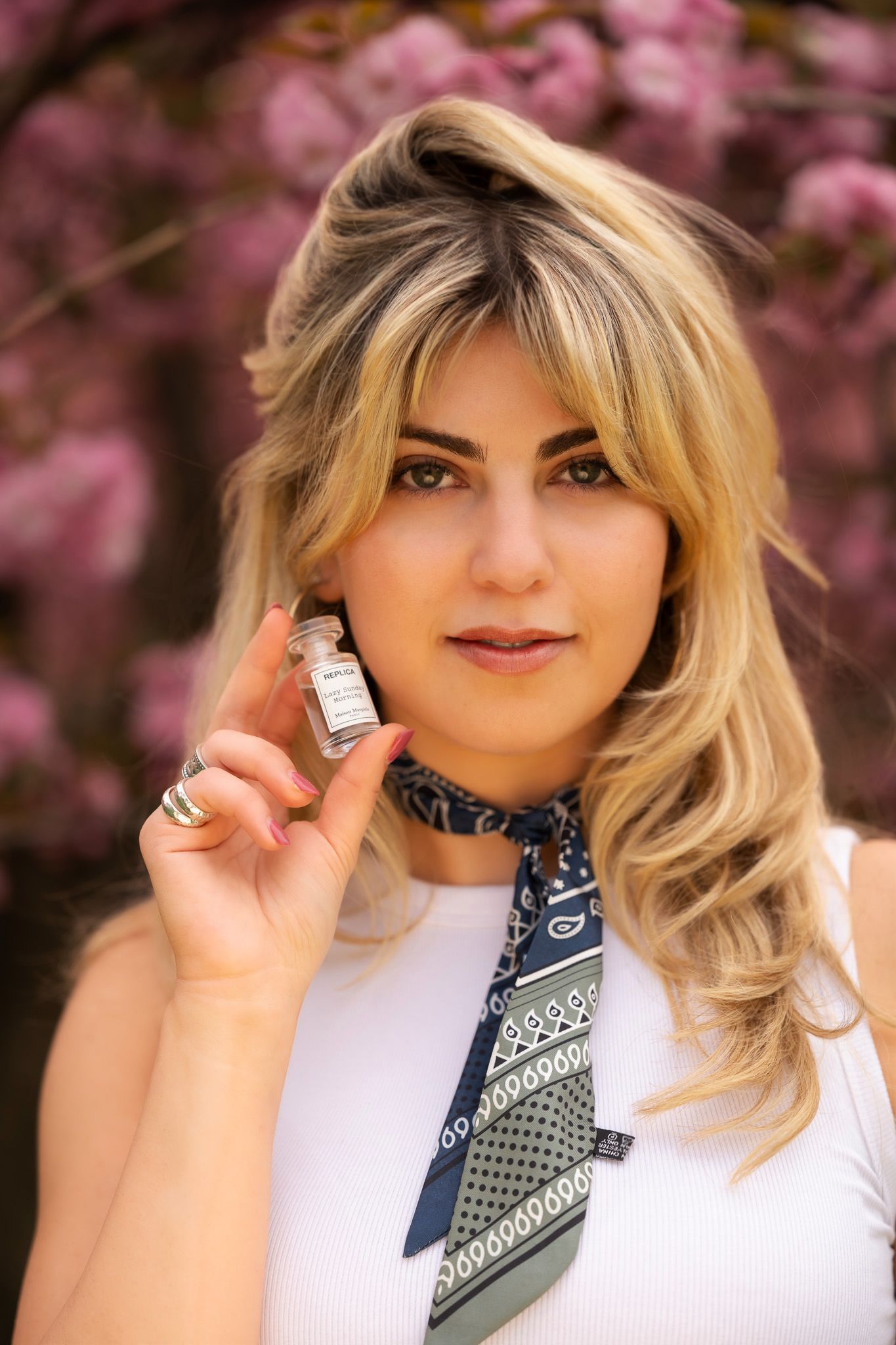 Blonde woman wearing white top and navy bandana scarf holding perfume bottle near pink blossoms.