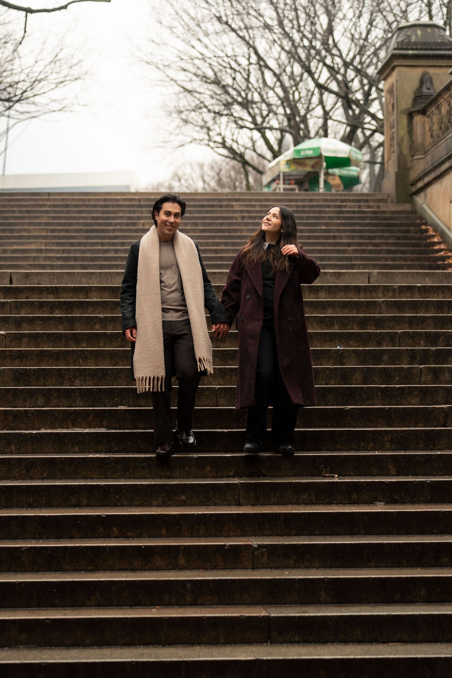 A couple stands together on stone steps in winter, wearing coats and scarves on a cloudy day.