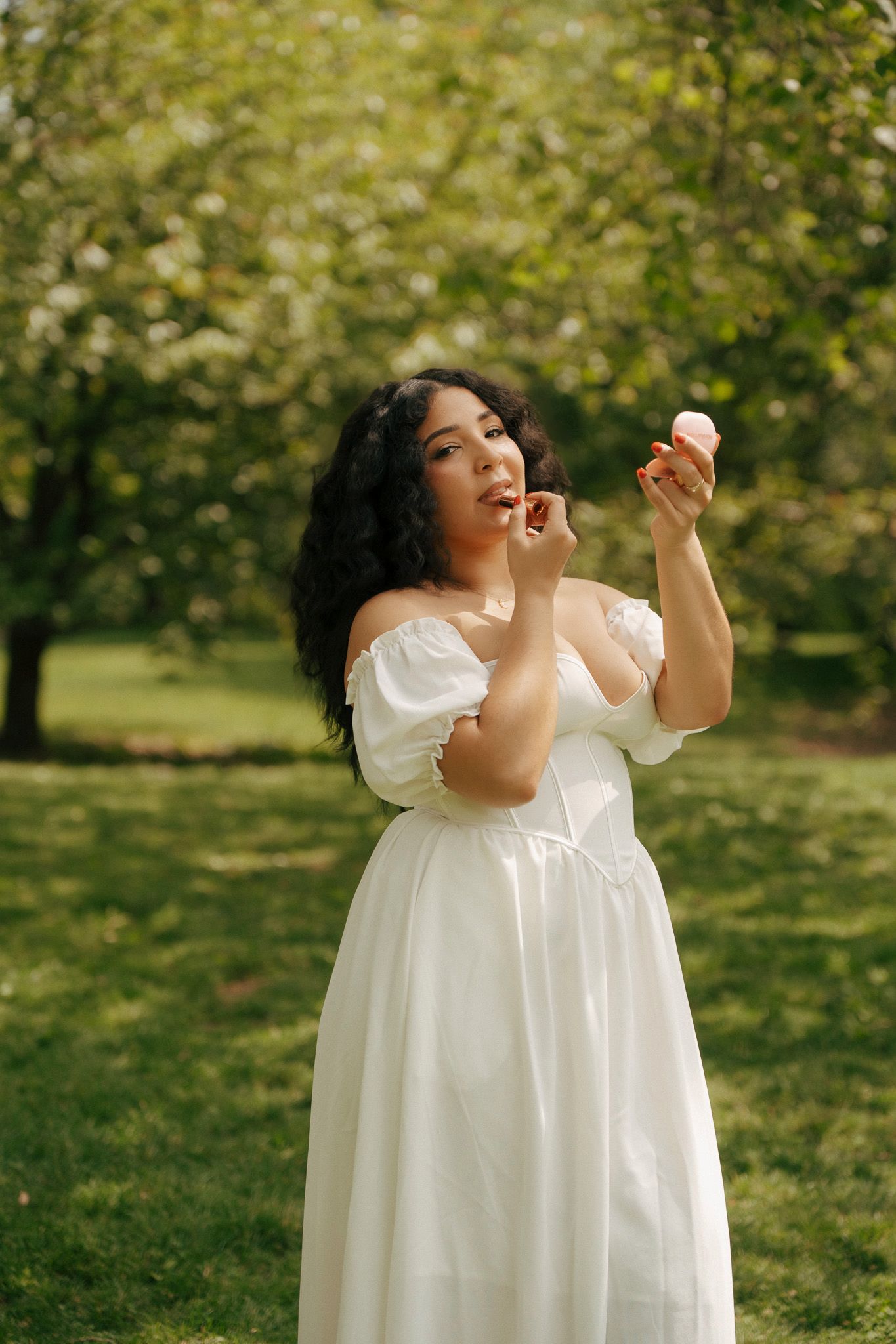 Woman in white off-shoulder dress eating a macaron in a lush green park setting.