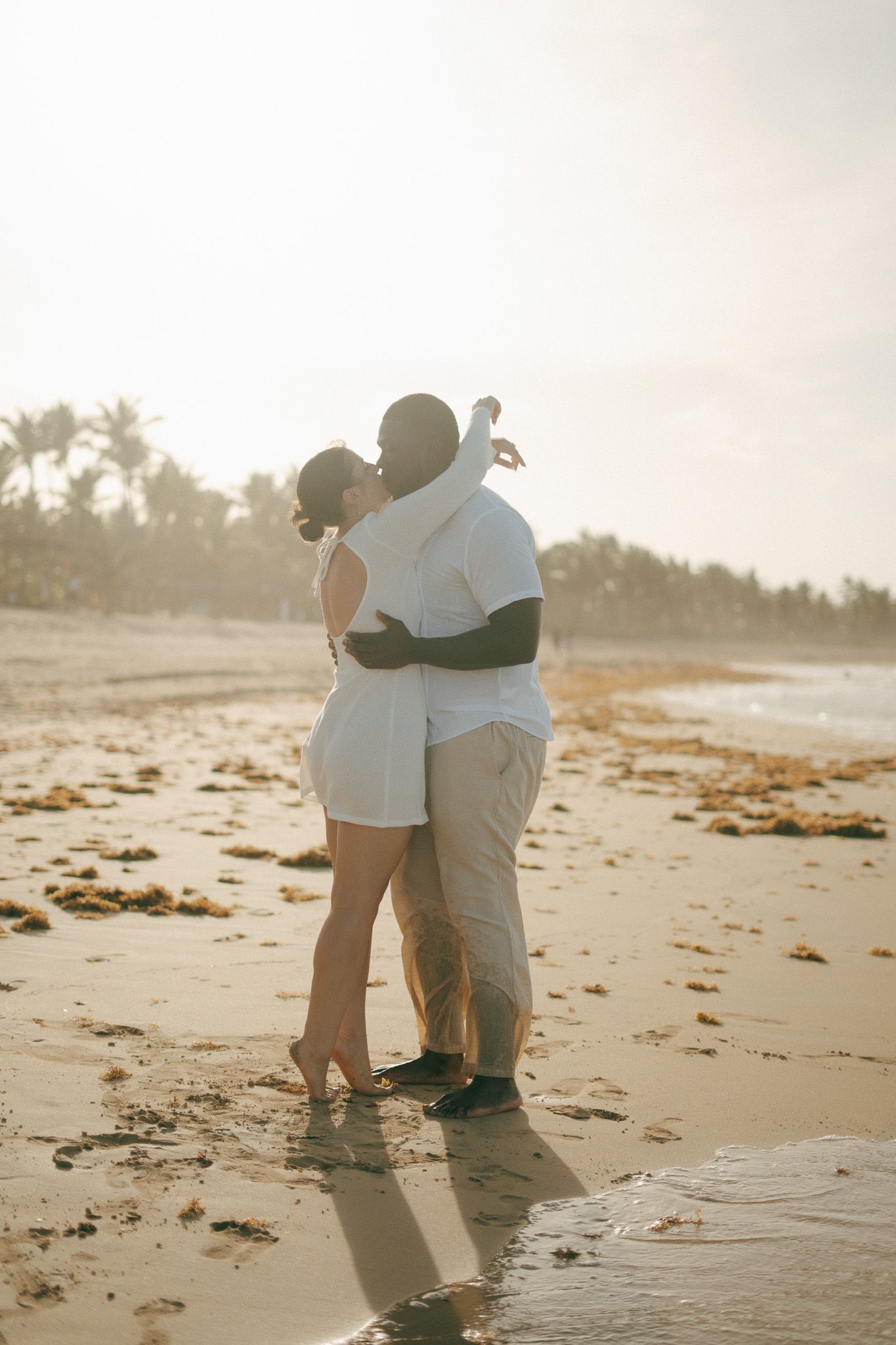 Couple sharing a romantic kiss on a sunlit beach, dressed in white, with palm trees and golden light in the background.
