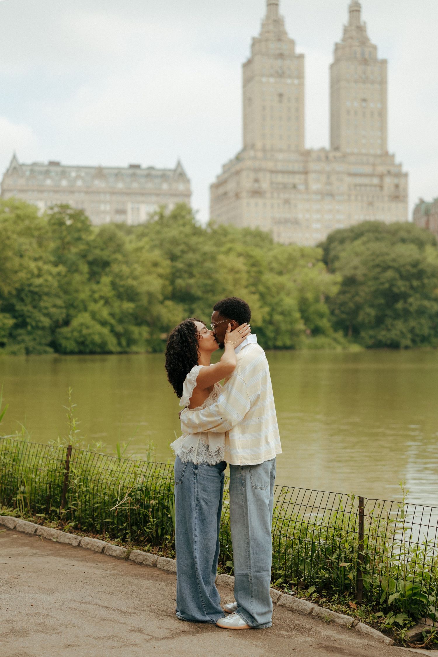 Romantic couple kissing by a lake in Central Park with iconic NYC buildings in the background.