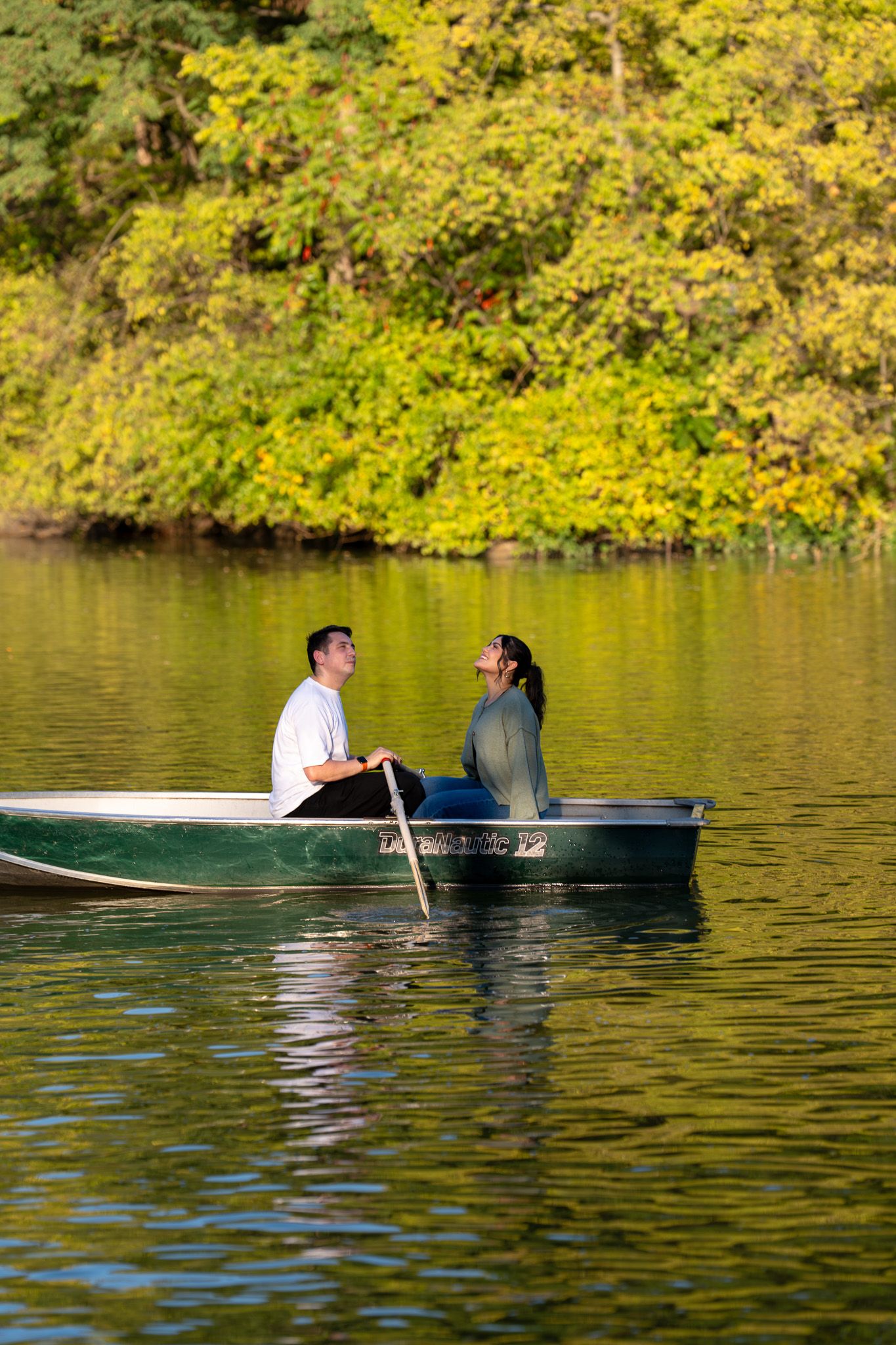 A couple rowing a boat across a lake surrounded by autumn foliage.