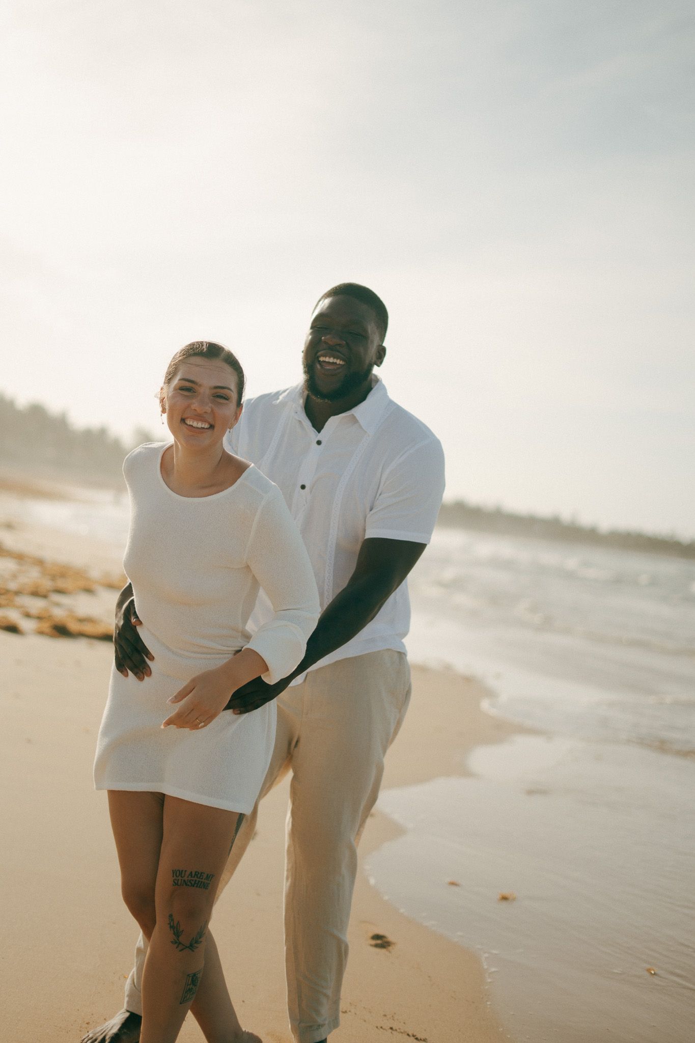 Happy interracial couple smiling and embracing on a sunny beach, wearing white outfits with ocean waves in the background.