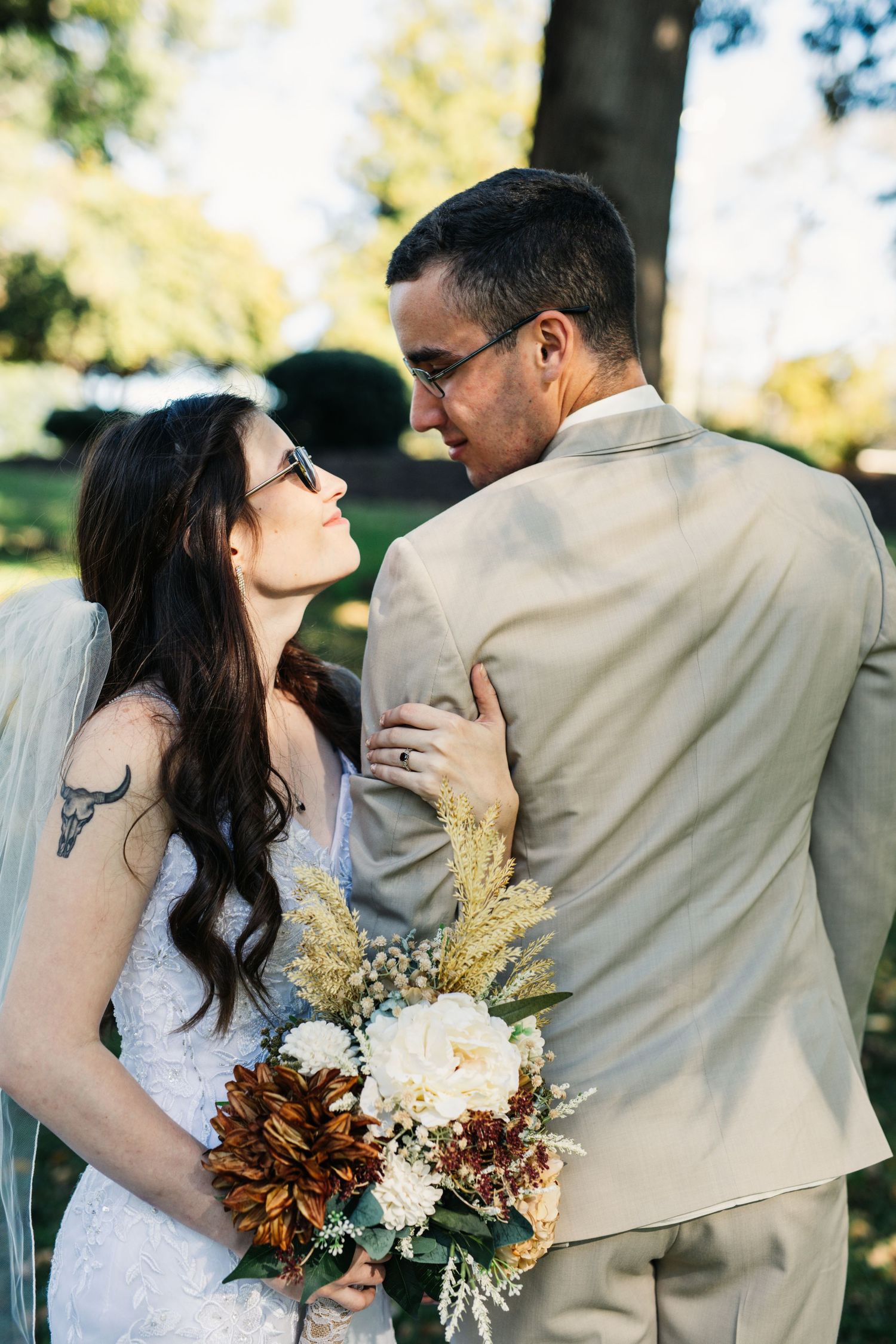 Bride holding autumn-themed bouquet gazes lovingly at groom in beige suit during outdoor wedding photoshoot.