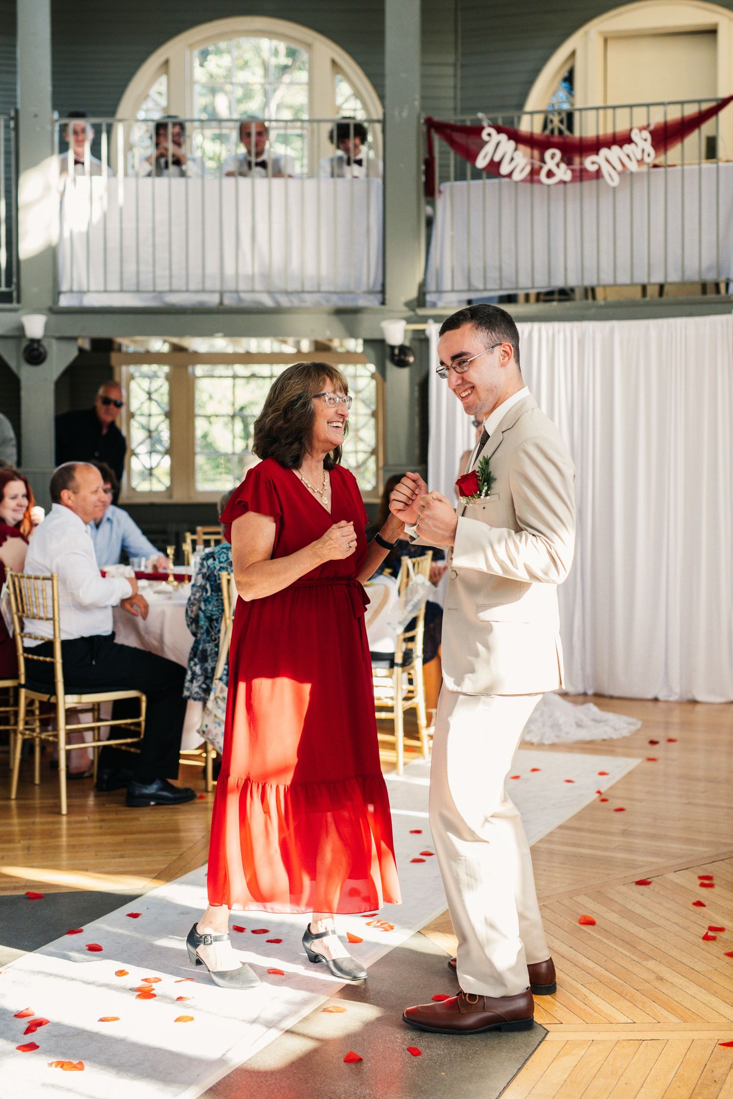 Wedding reception features couple dancing on wooden floor with red decorations and arched windows.