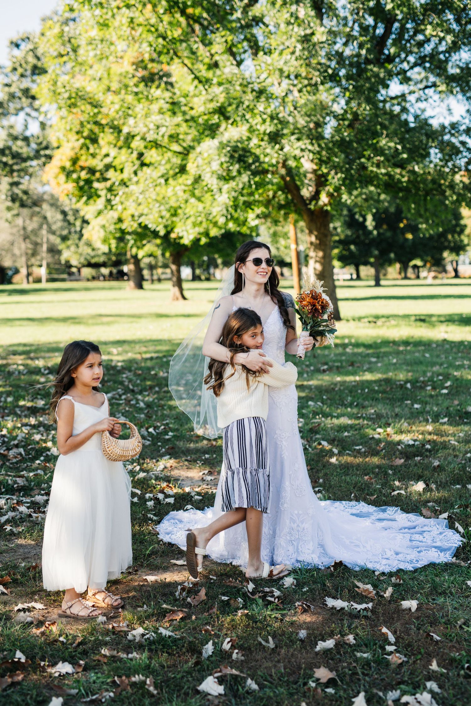 Wedding party members gather under trees in a park with sunlight filtering through the branches.