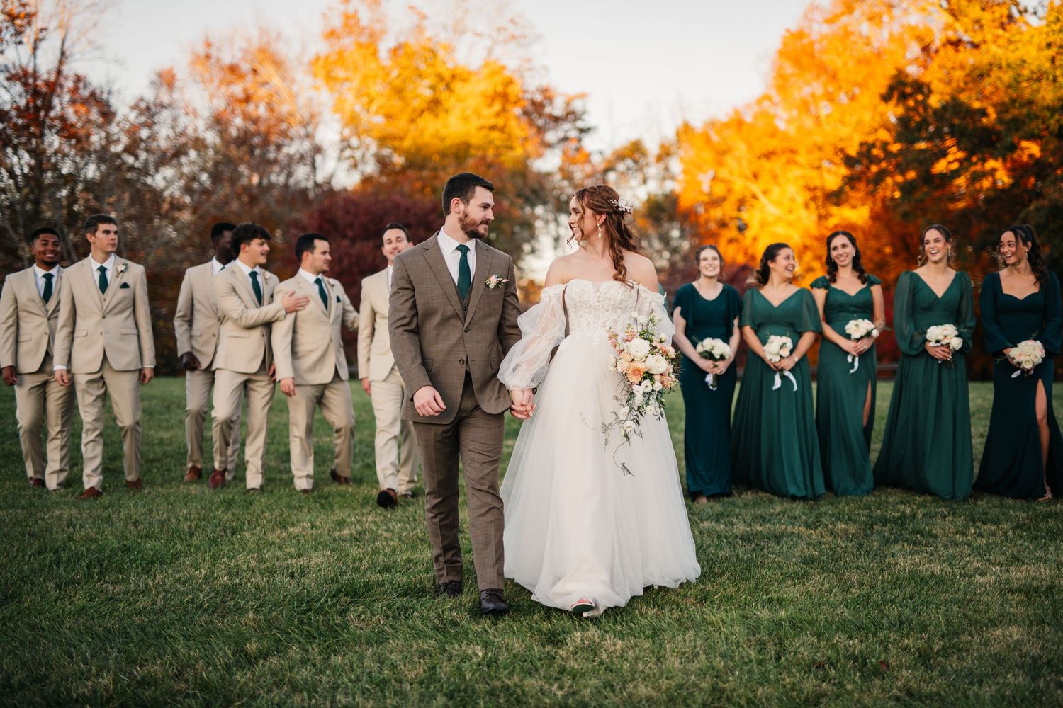 A fall wedding party poses on grass with vibrant orange foliage behind them.