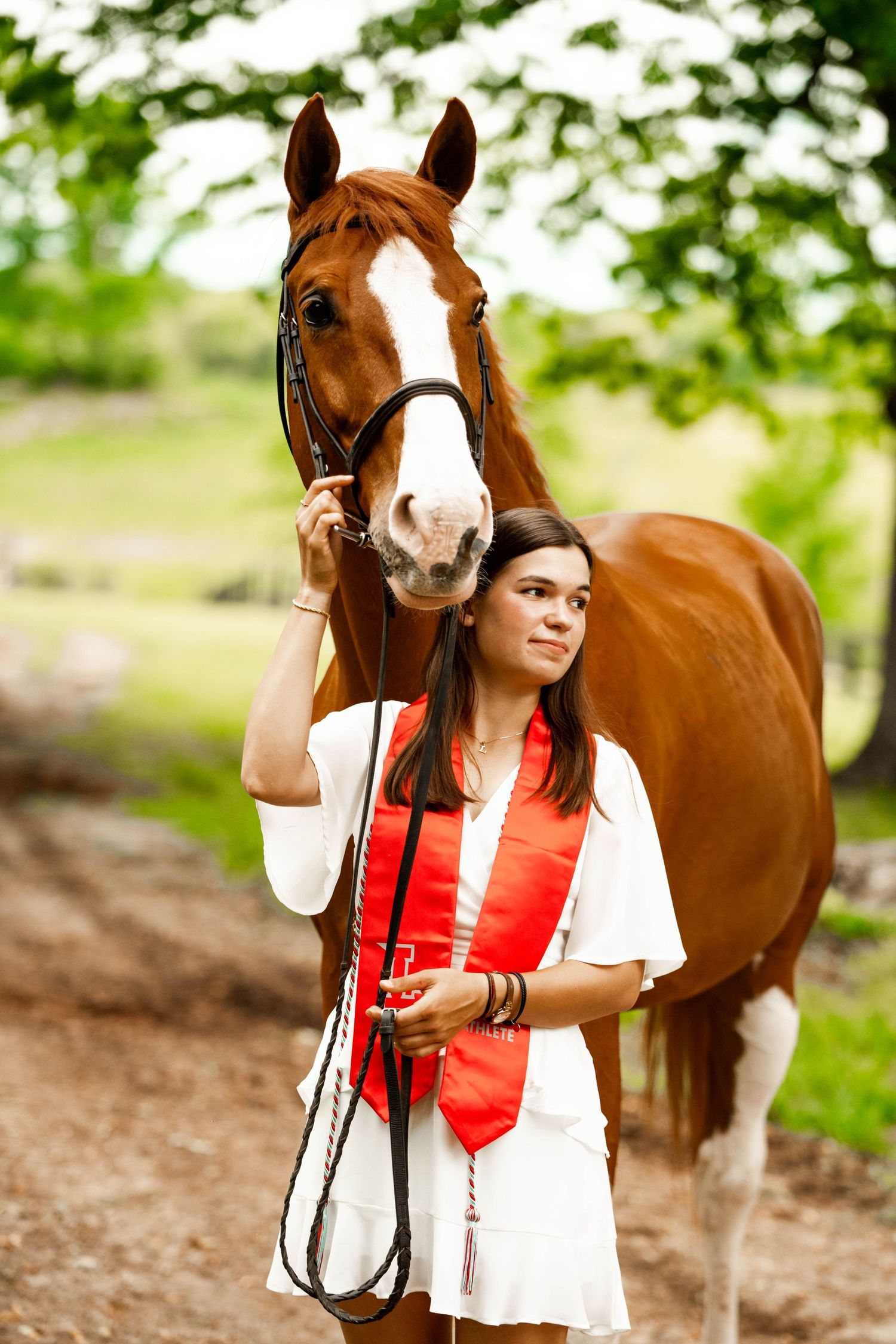 A chestnut horse with a white blaze stands with someone wearing graduation attire on a wooded path.