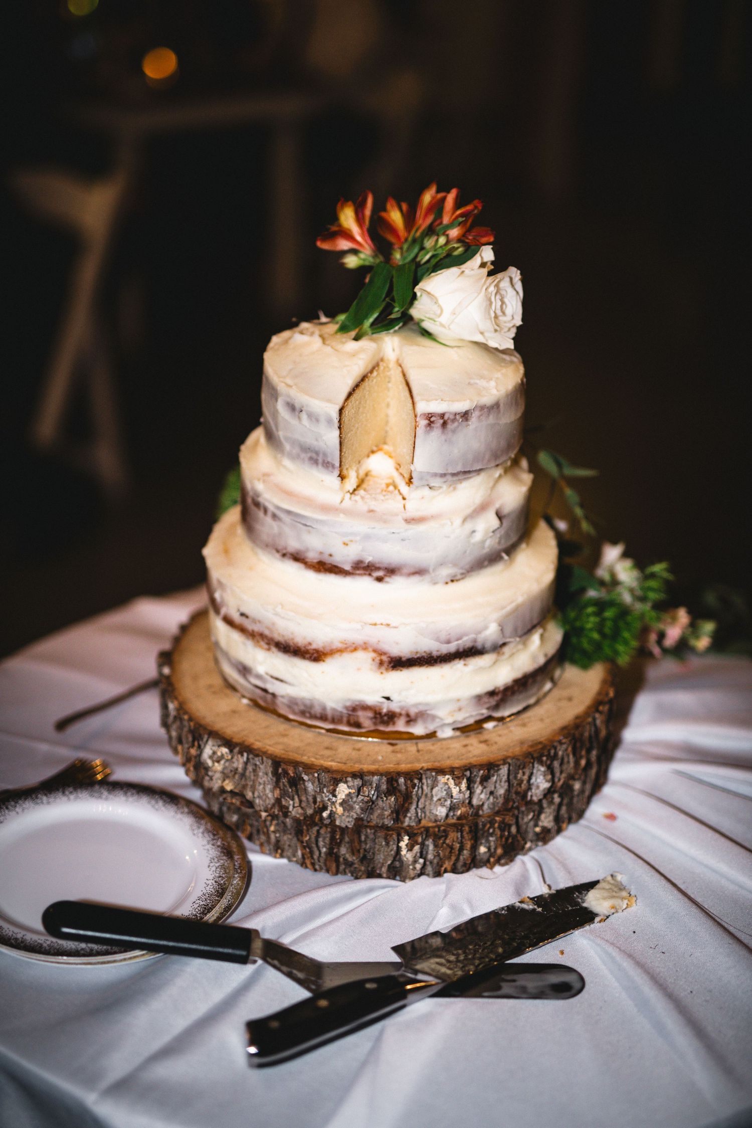 A semi-naked wedding cake decorated with fresh flowers sits on a wooden slice cake stand.