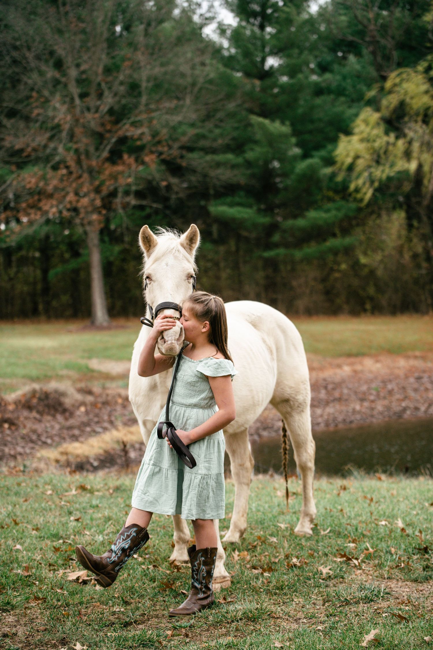A white horse and someone in a light blue dress pose together on an autumn afternoon in a wooded area.