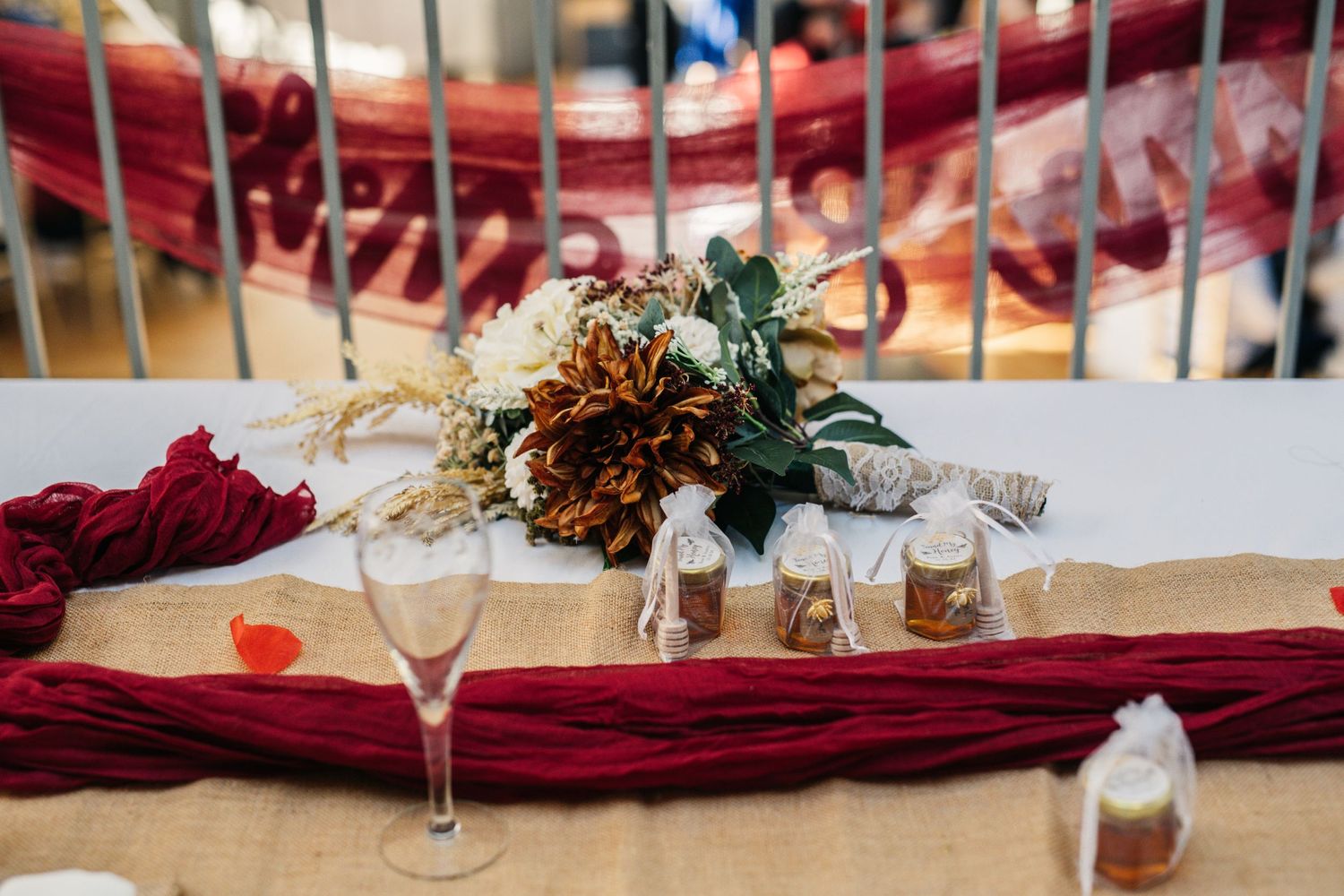Wedding reception table with burgundy runner and floral centerpiece against red chairs.