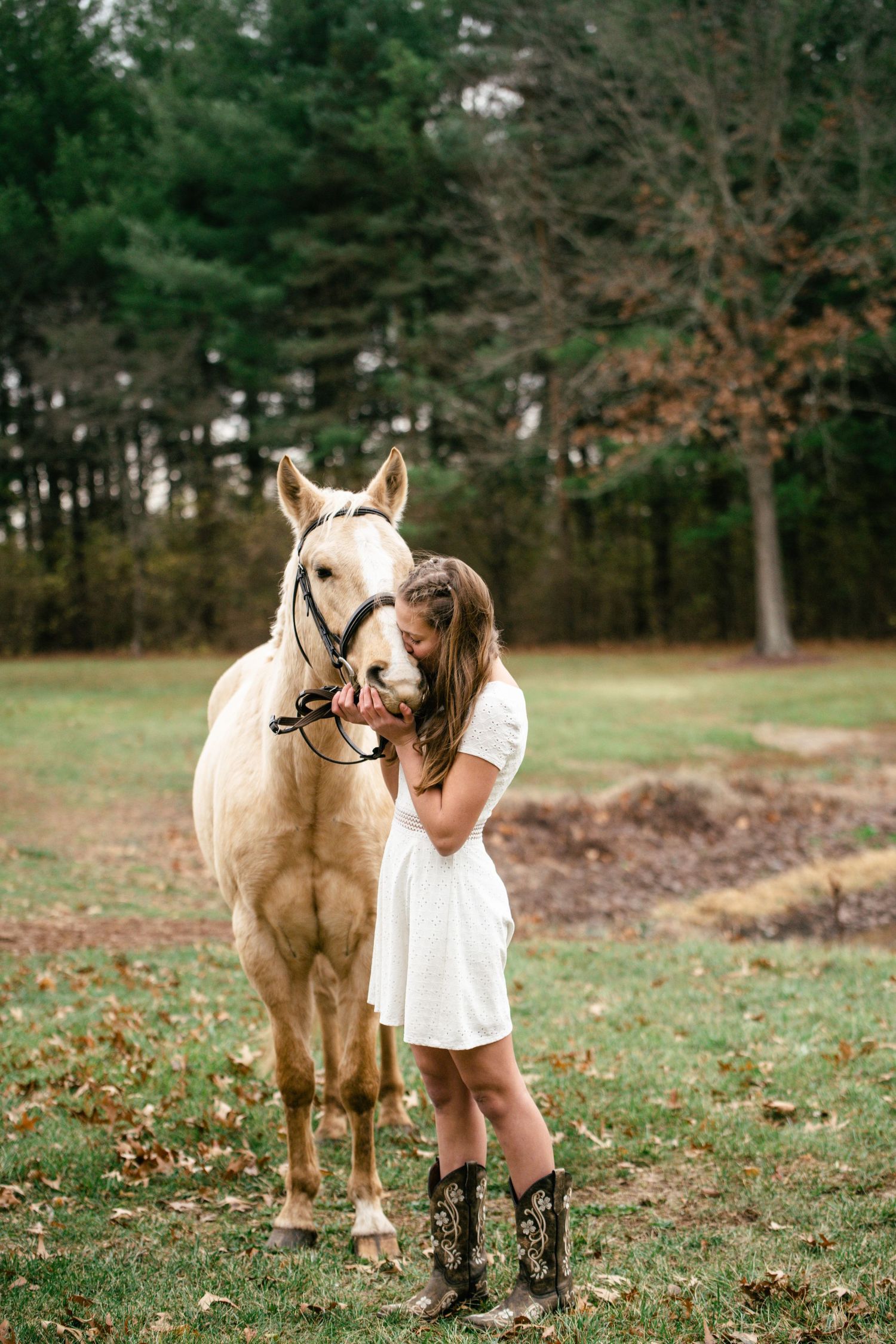 Person in white dress and boots embraces white horse in rustic outdoor setting.