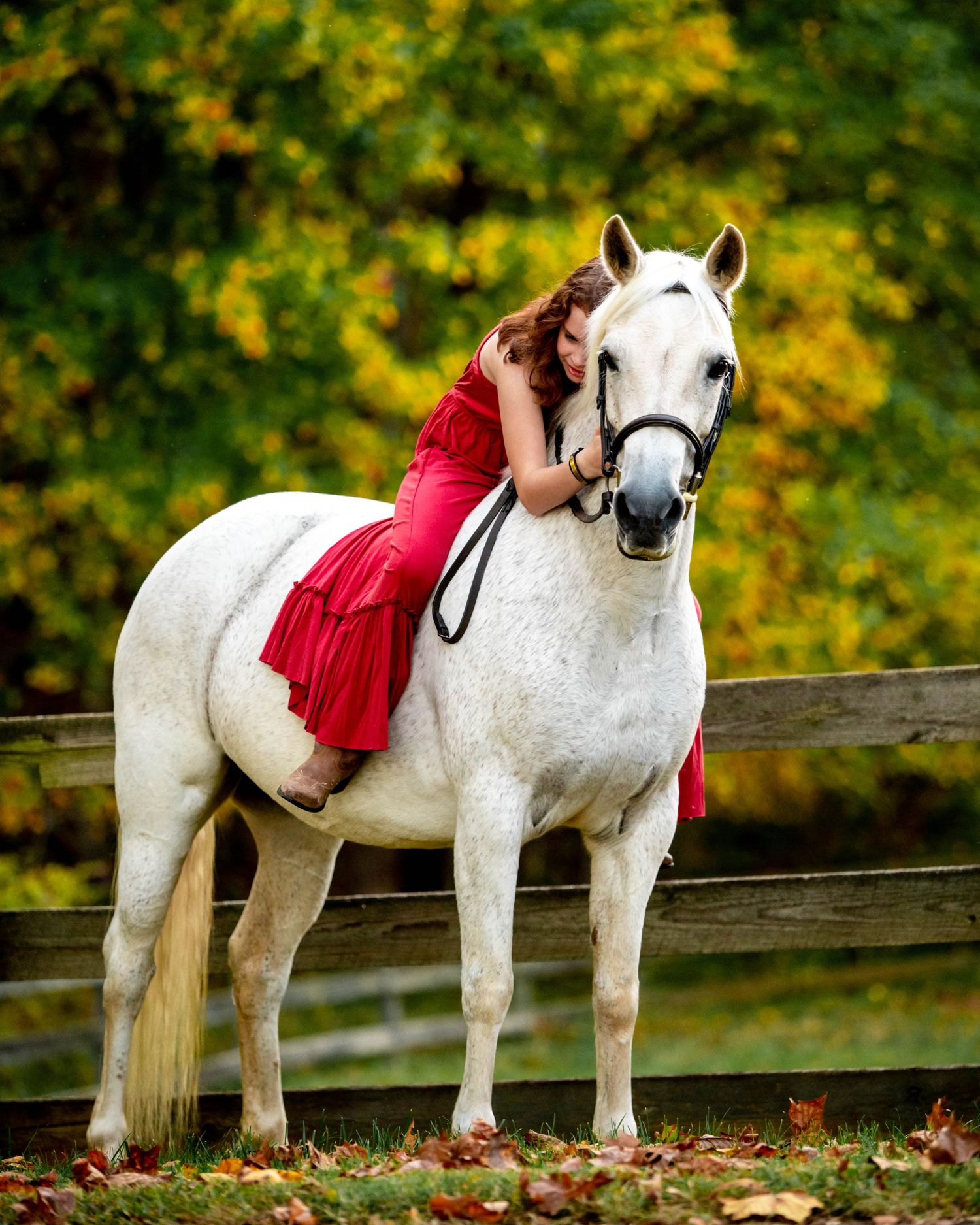 A white horse and rider in a red dress pose together against colorful fall foliage.