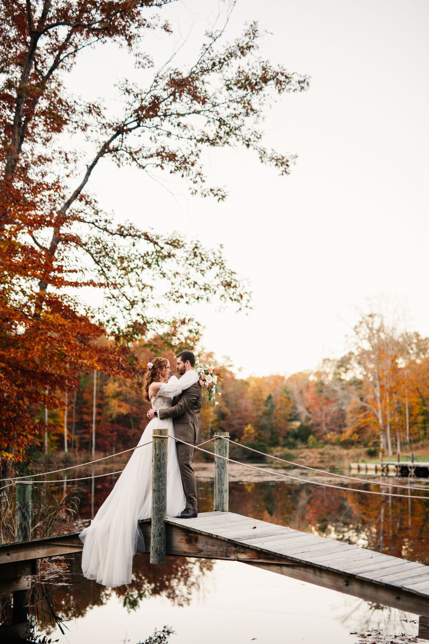 A romantic wedding couple embraces on a wooden bridge surrounded by autumn colors.