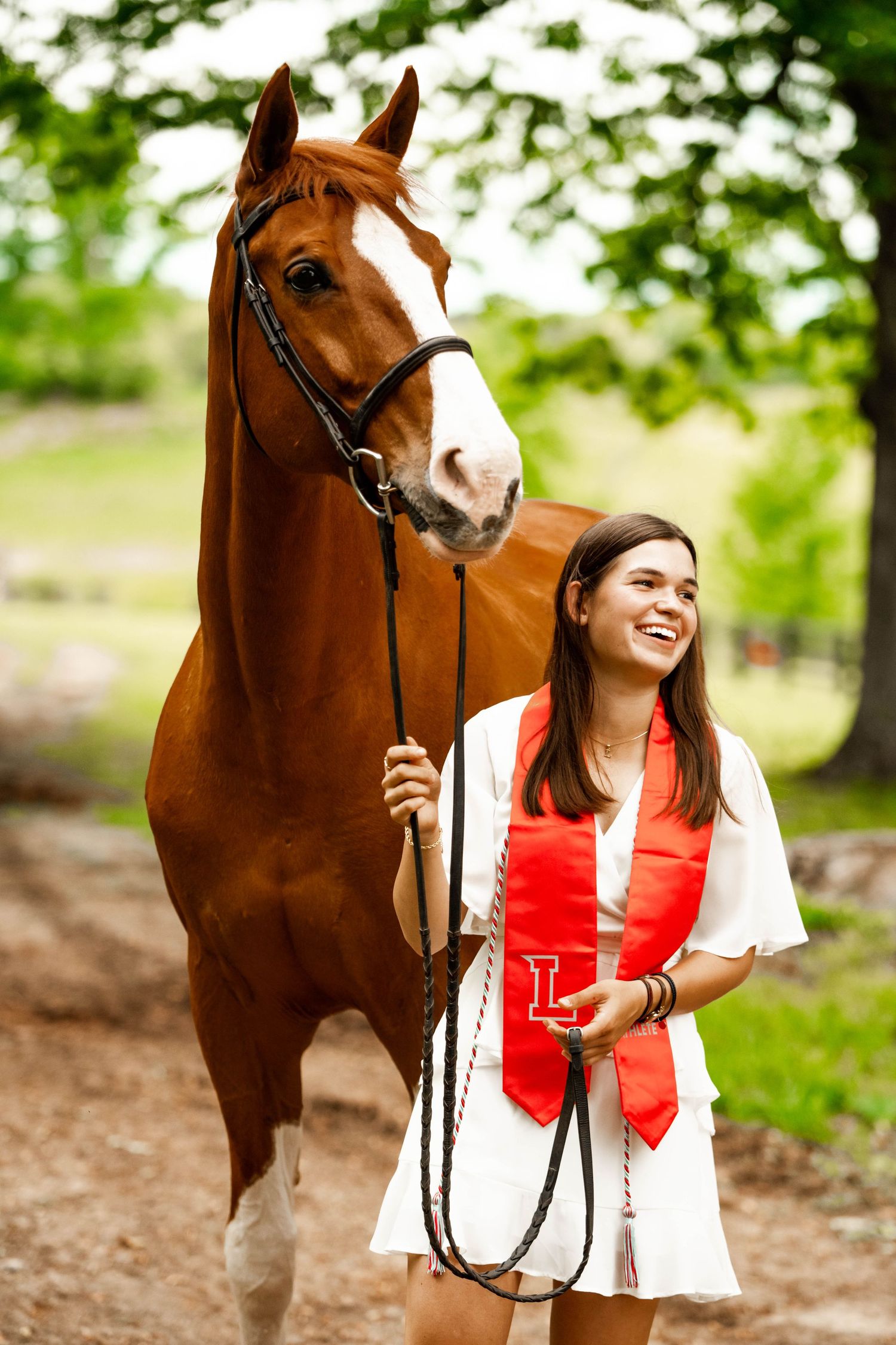 A chestnut horse and graduate in white dress with red sash pose together on a forest trail.