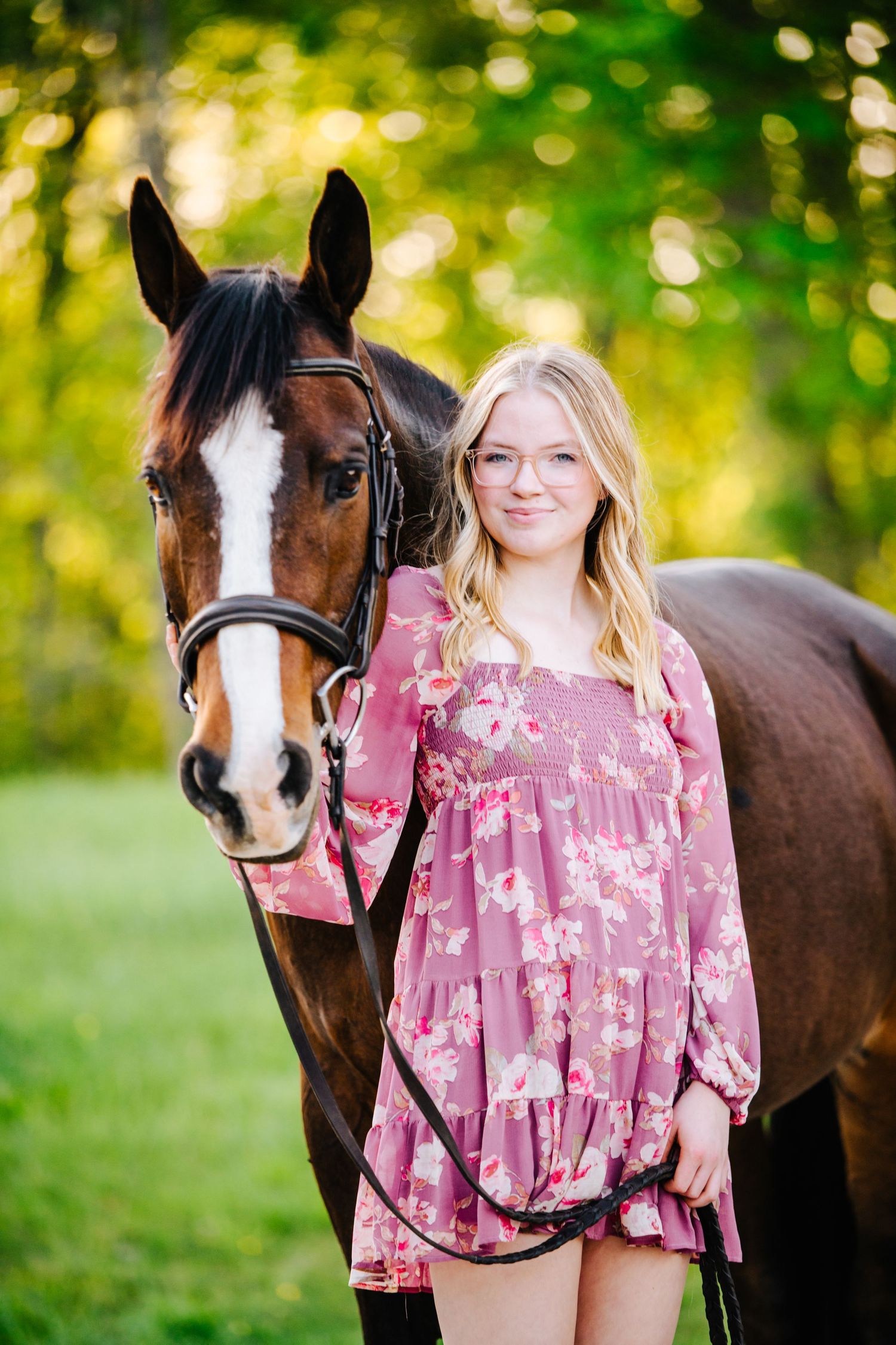 Bay horse with white blaze poses with person wearing pink floral dress in sunlit field.