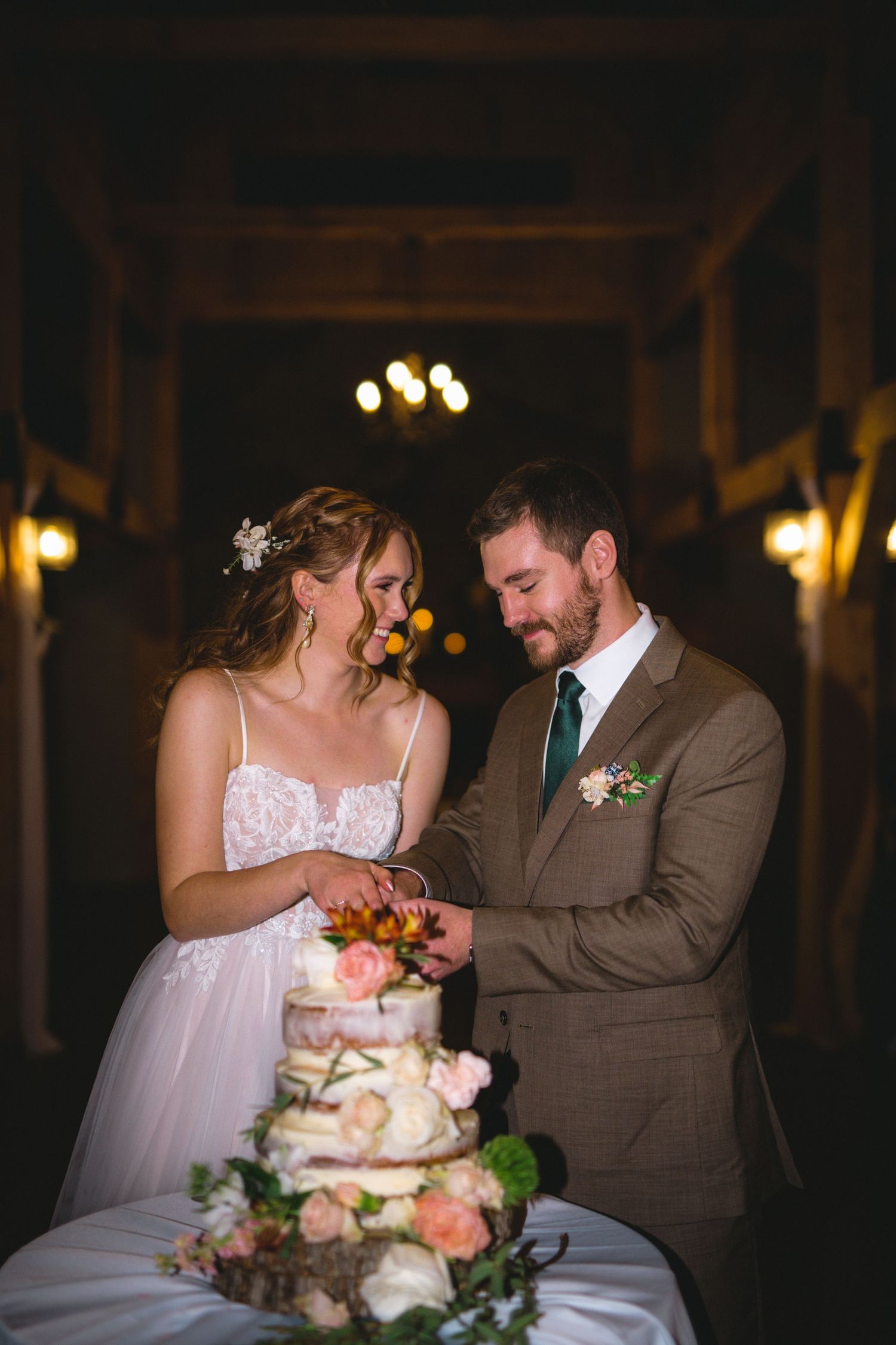 A couple cuts their rustic wedding cake decorated with fresh flowers in warm candlelight.
