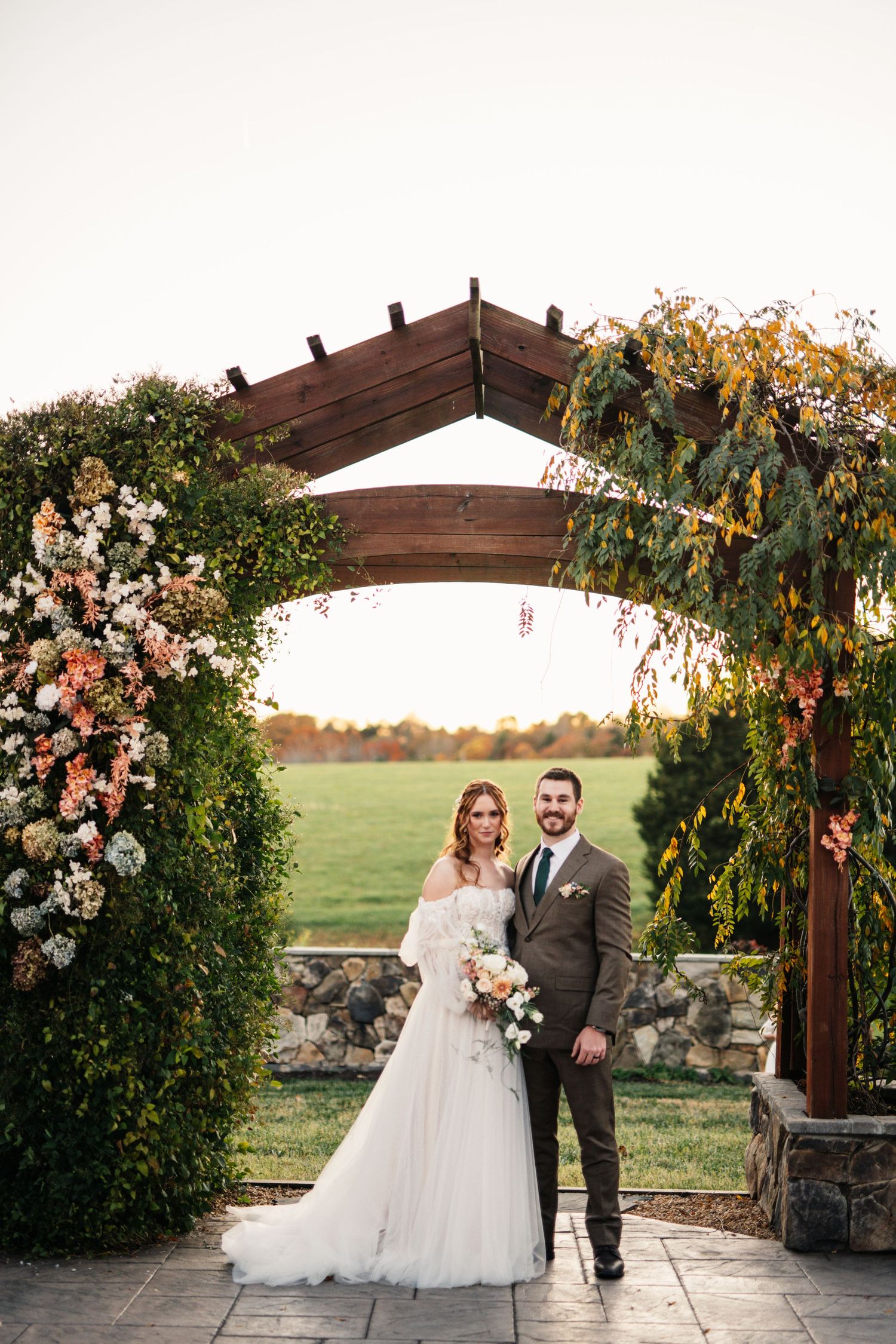 Wedding couple poses under floral arch with stone base during golden hour.