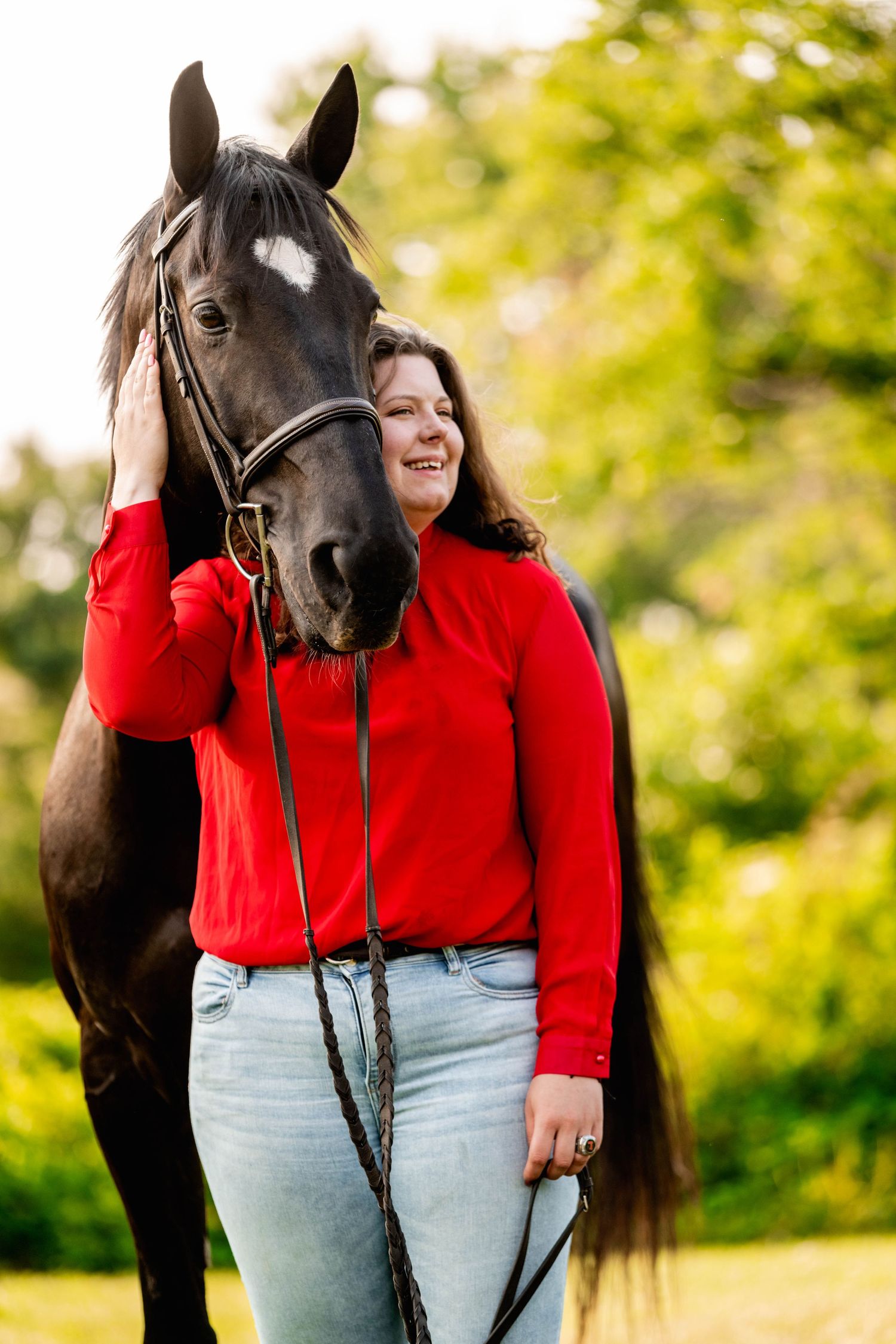 A dark bay horse with bridle stands next to someone in a bright red sweater on a sunny day.