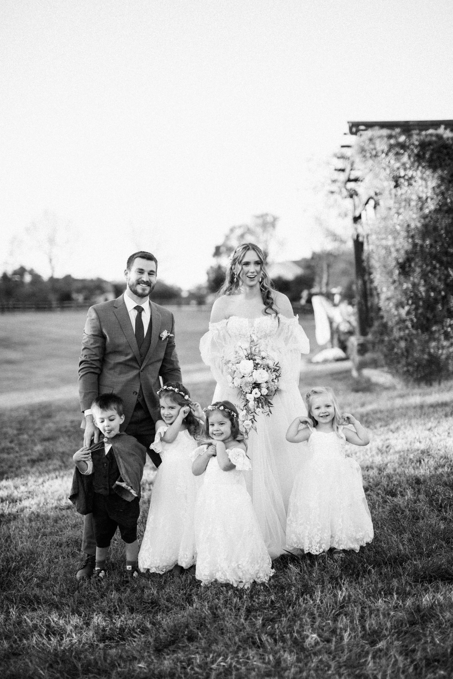 A wedding couple poses with three young flower girls in white dresses on a grassy hillside.
