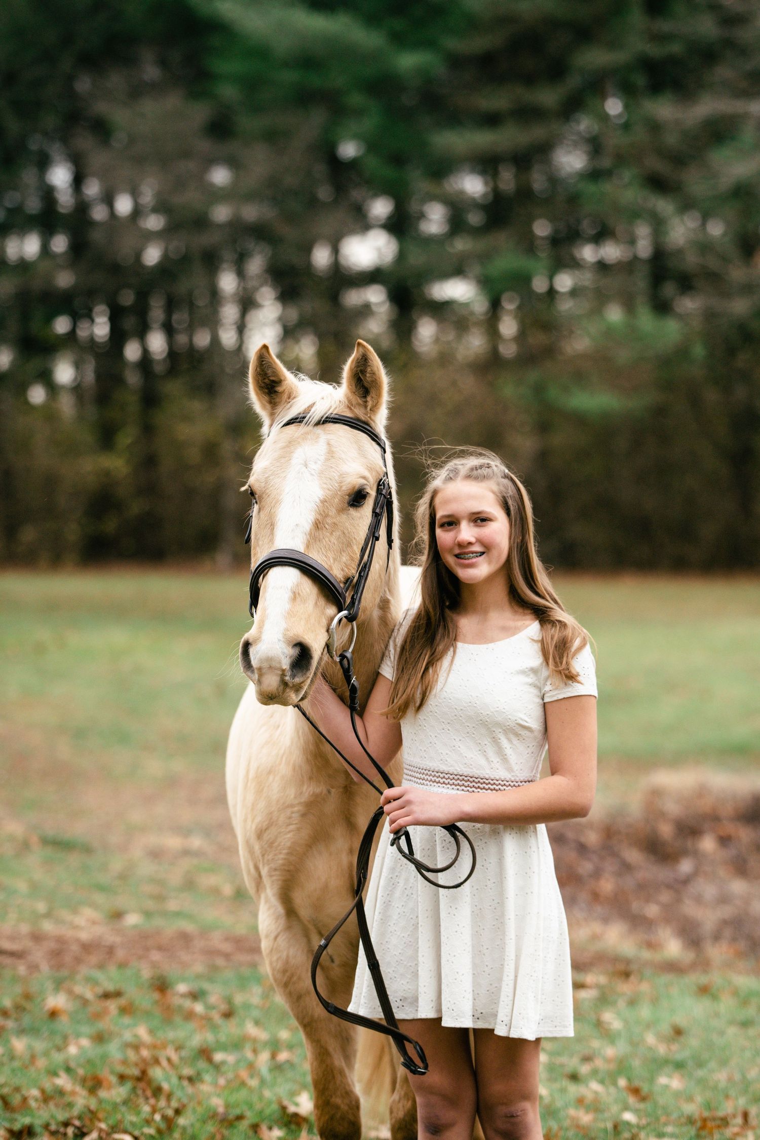 Palomino horse and person in white dress pose together in front of pine trees.