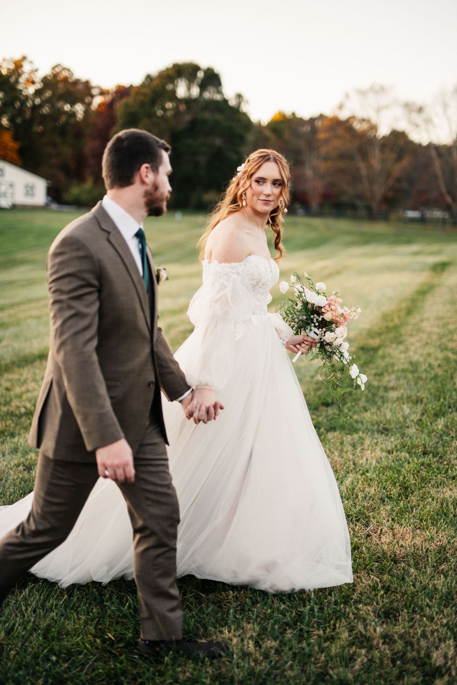 A couple in wedding attire strolls through a scenic fall meadow.