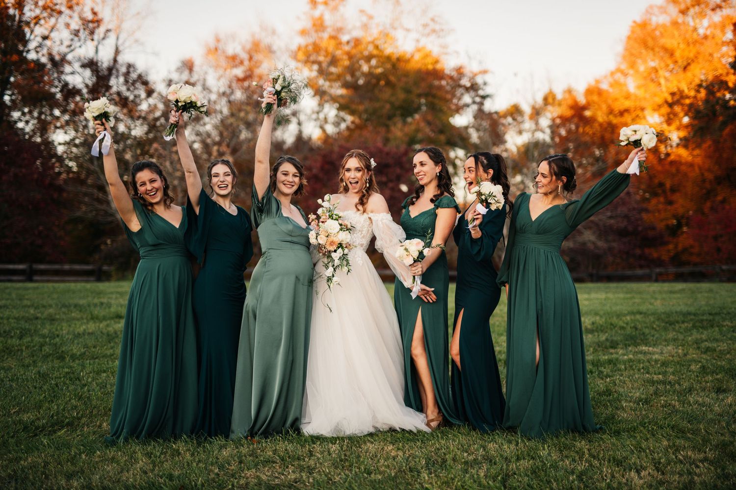 Bridal party in emerald green dresses pose with bride against autumn sunset backdrop.