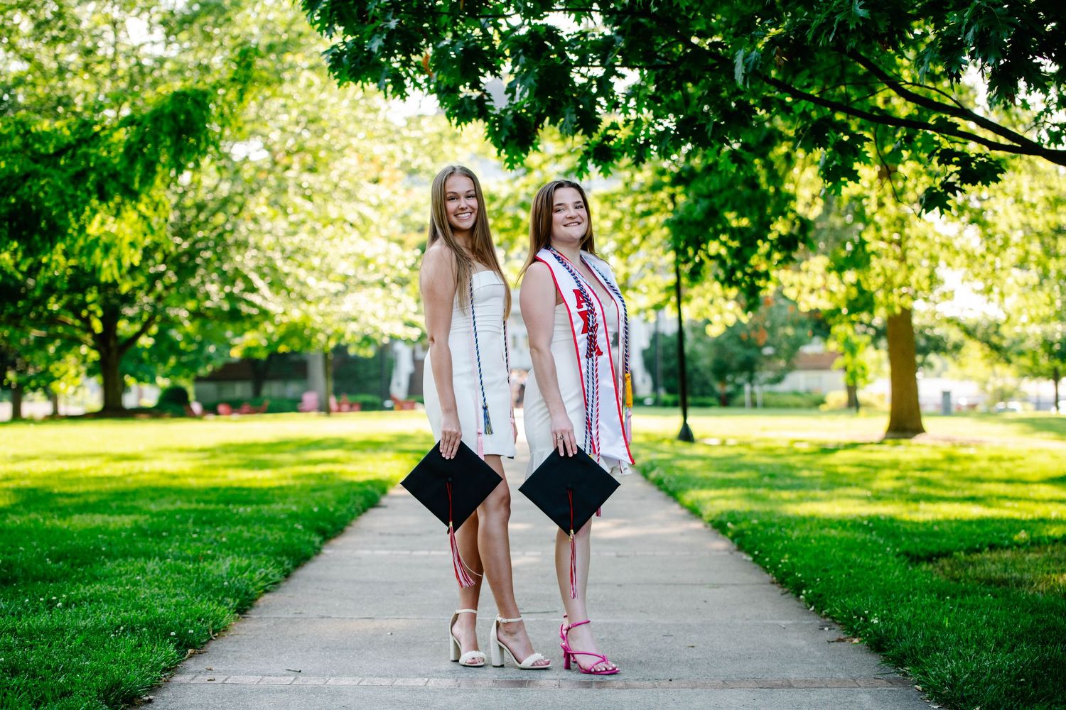 Two graduates in white dresses pose on a tree-lined campus walkway.