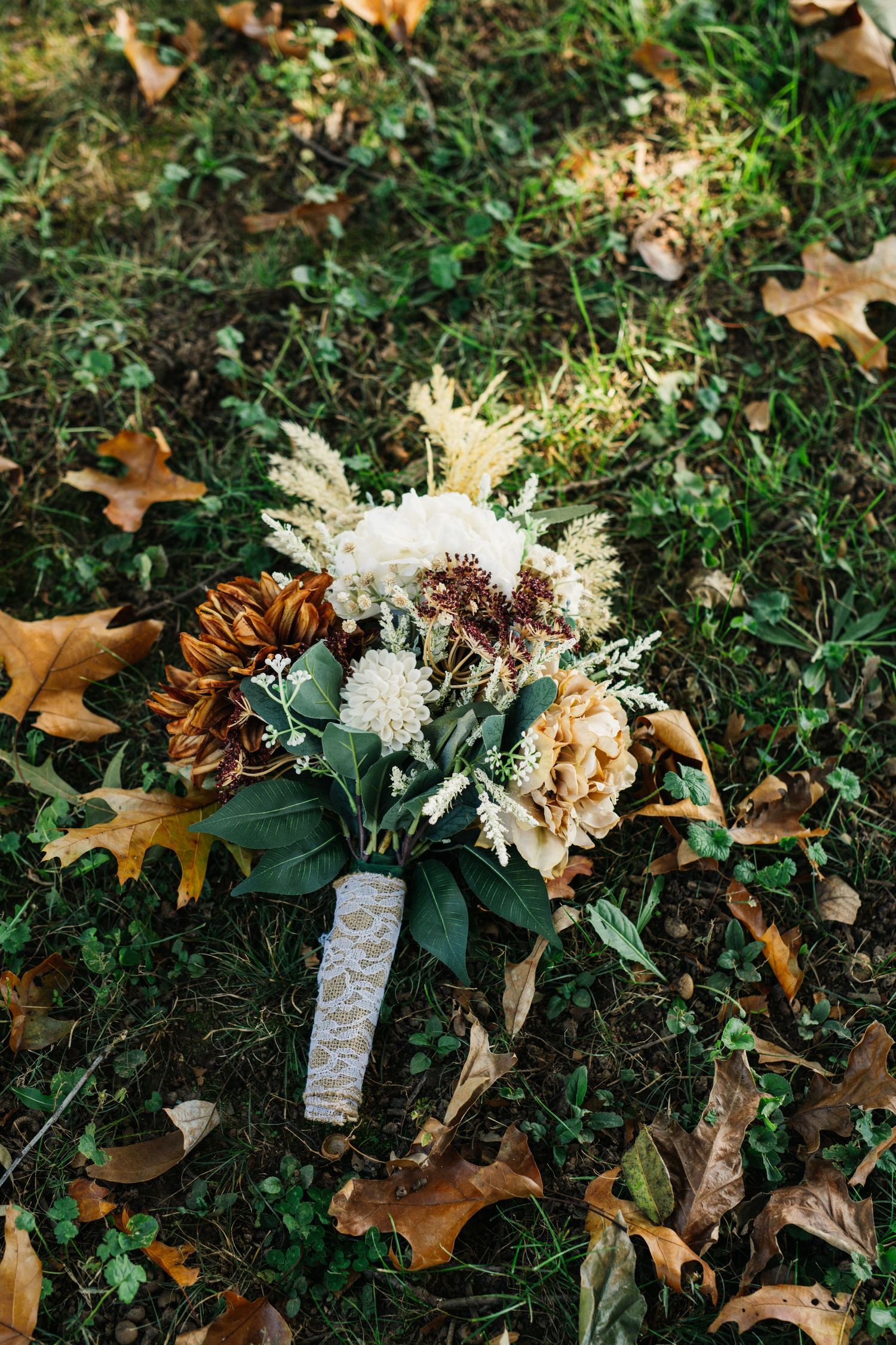 Wedding bouquet with white and brown flowers lying on grass surrounded by autumn leaves.