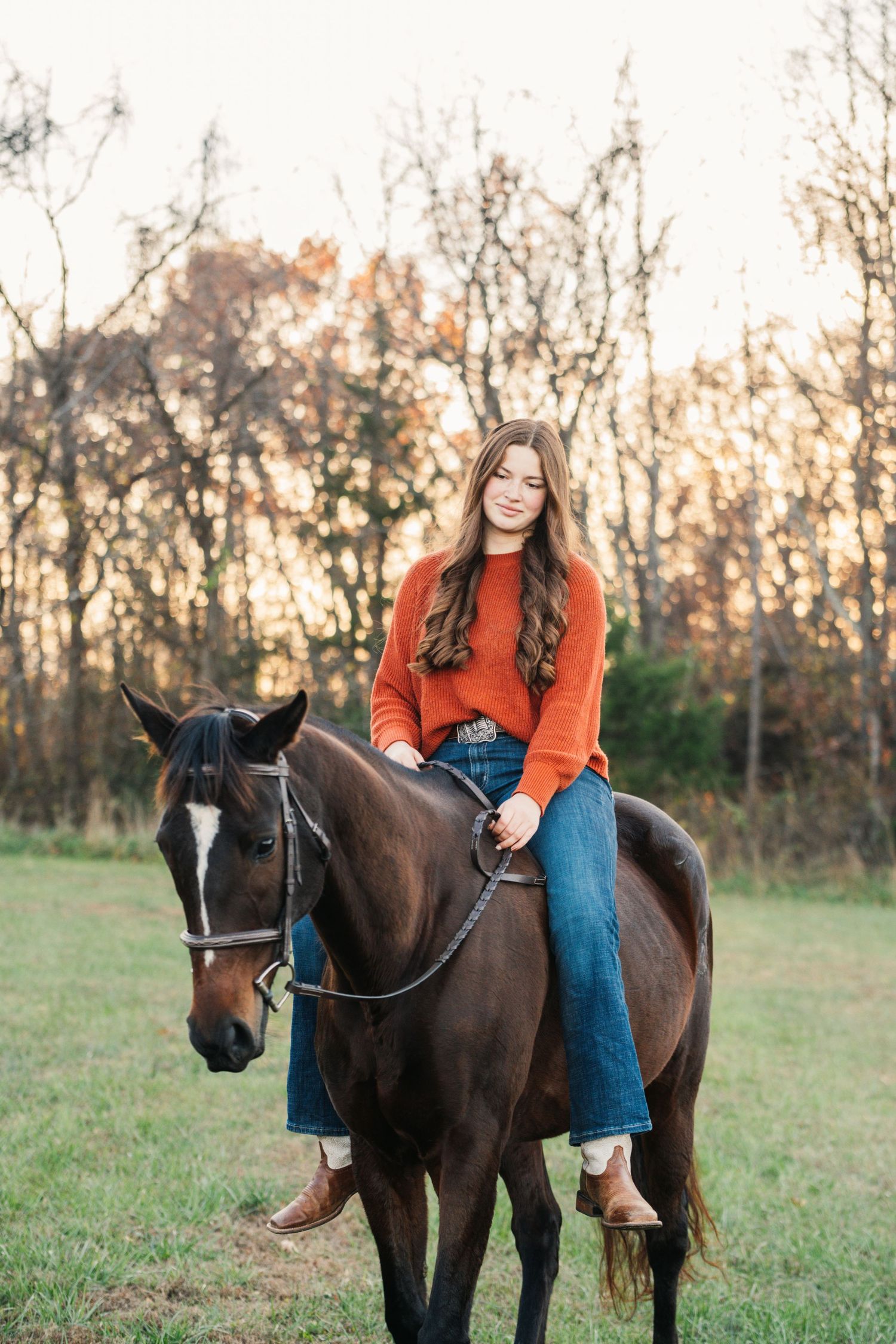 Person in bright orange sweater rides a dark bay horse in an autumn field at sunset.