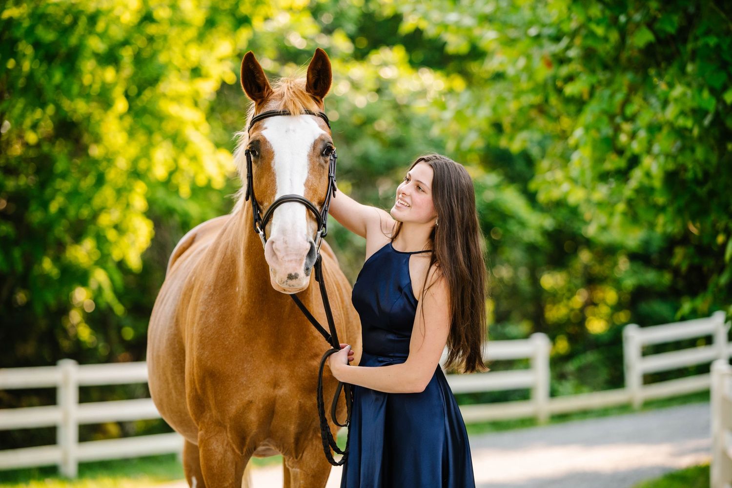 A chestnut horse with white markings stands beside companion in navy dress on wooded path.