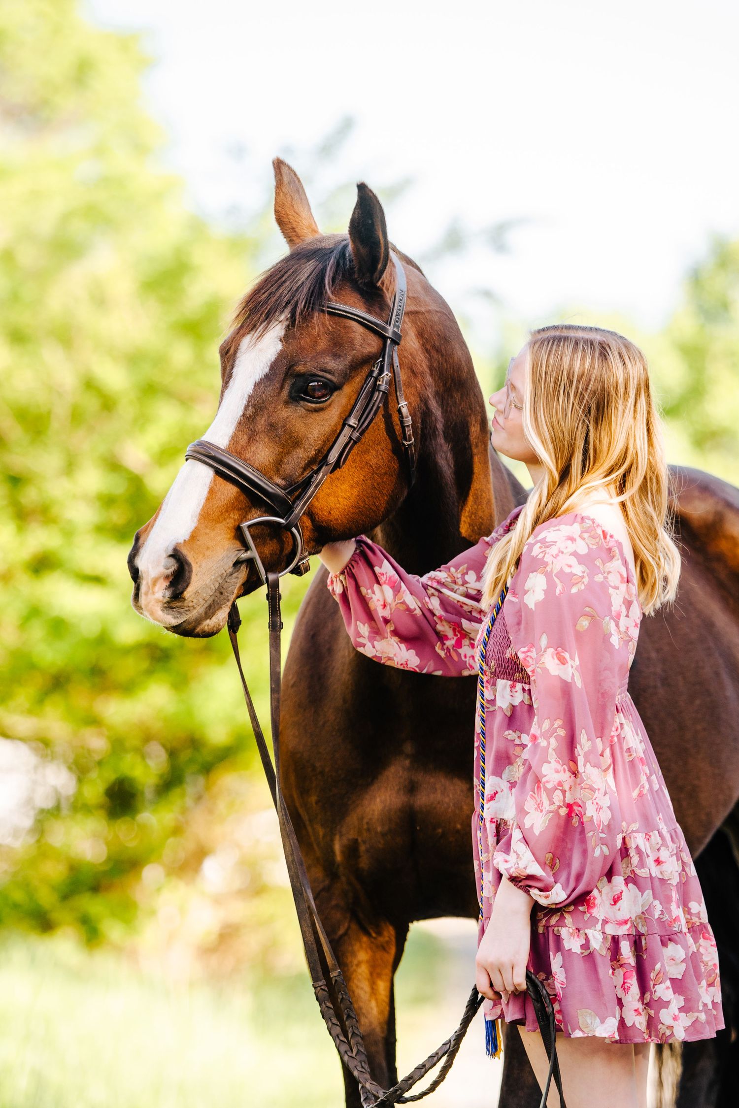 Bay horse with white blaze stands beside person in pink floral dress on sunny day.