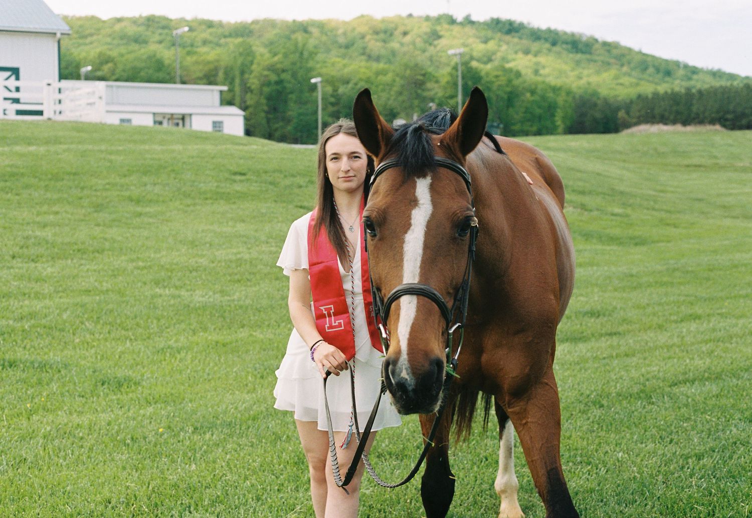 Student in white dress stands with bay horse wearing red sash against rural backdrop.