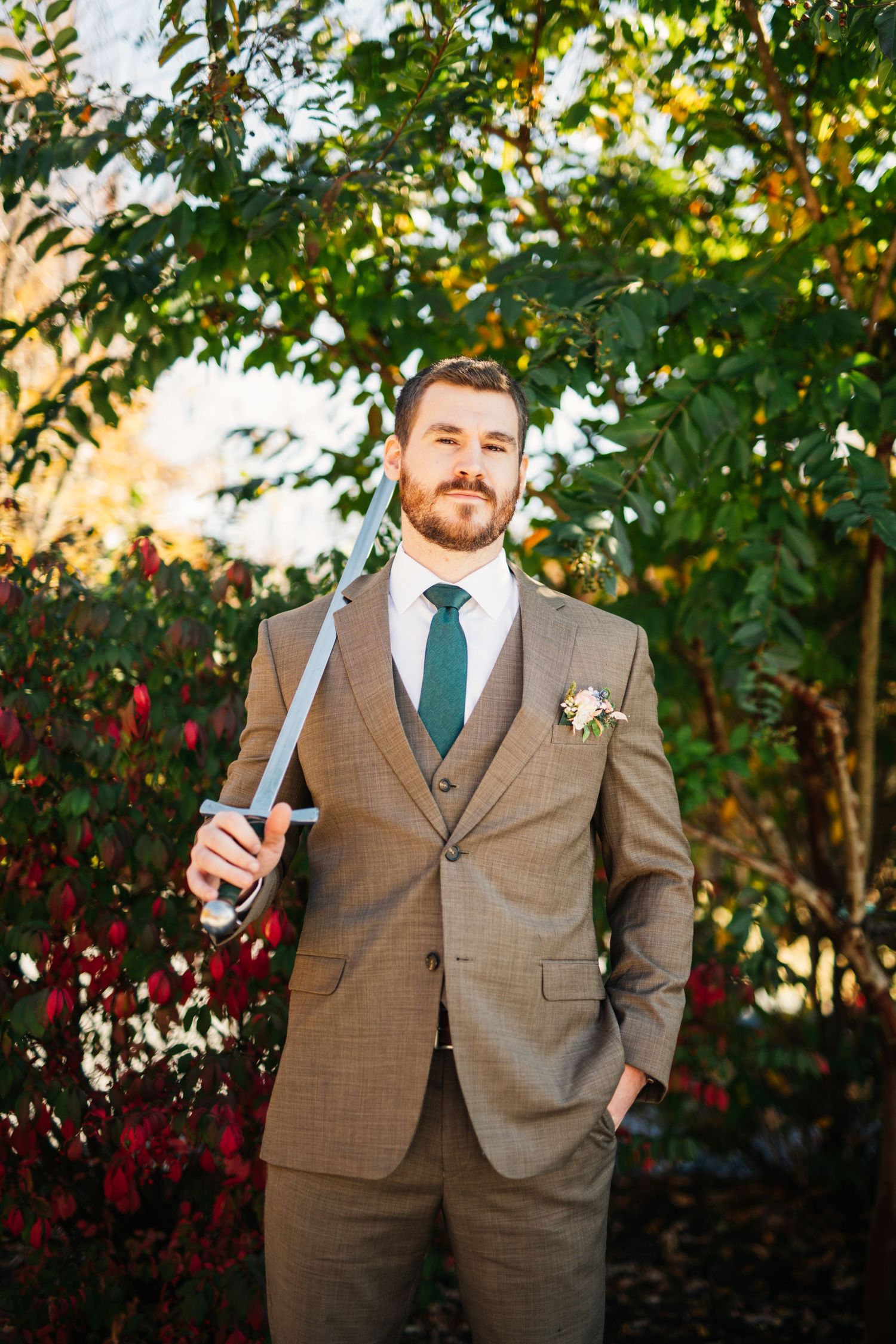 A groom in brown suit and green tie poses in garden setting during golden hour.