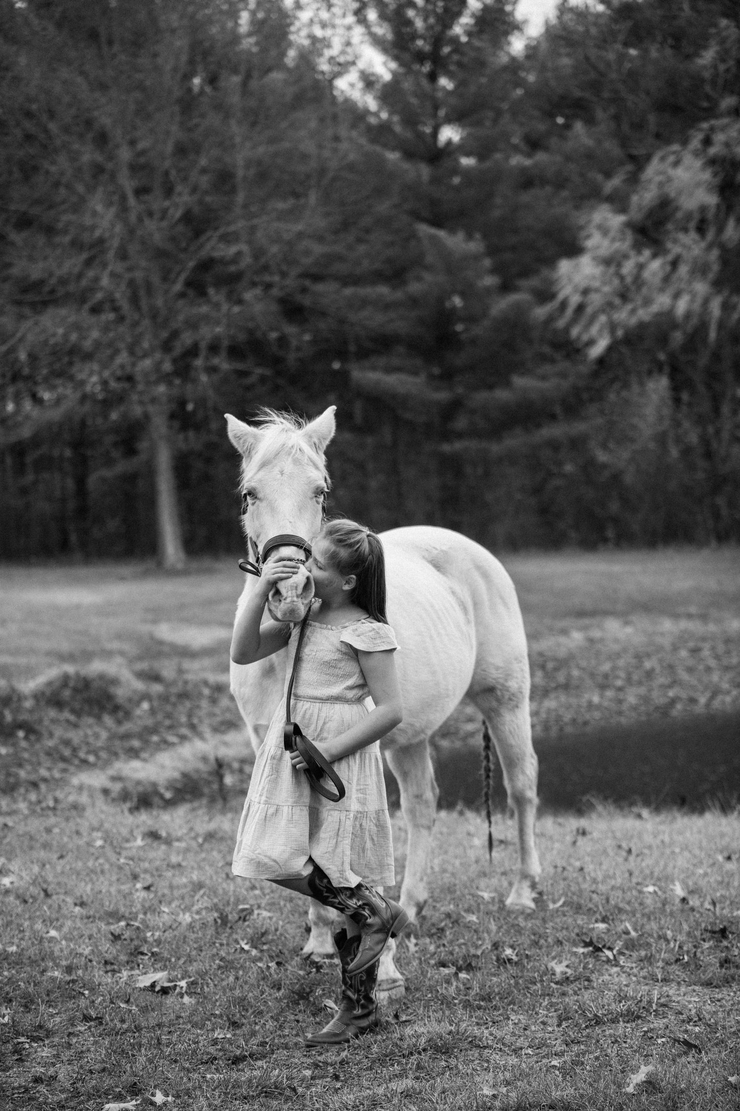 Black and white portrait of a white horse and companion in a dress and boots in a natural setting.
