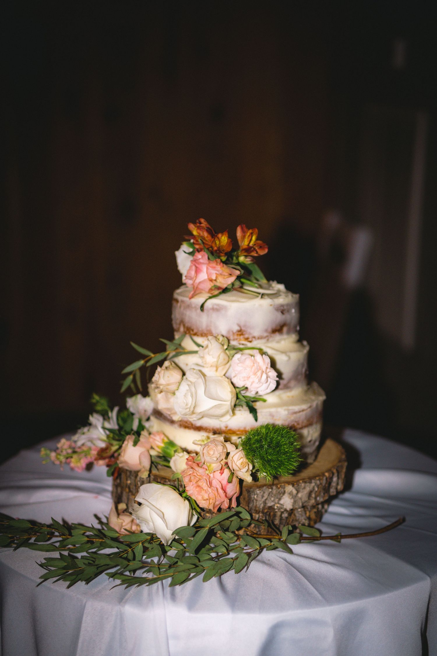 A three-tiered naked wedding cake decorated with fresh flowers and greenery on a wood slice stand.