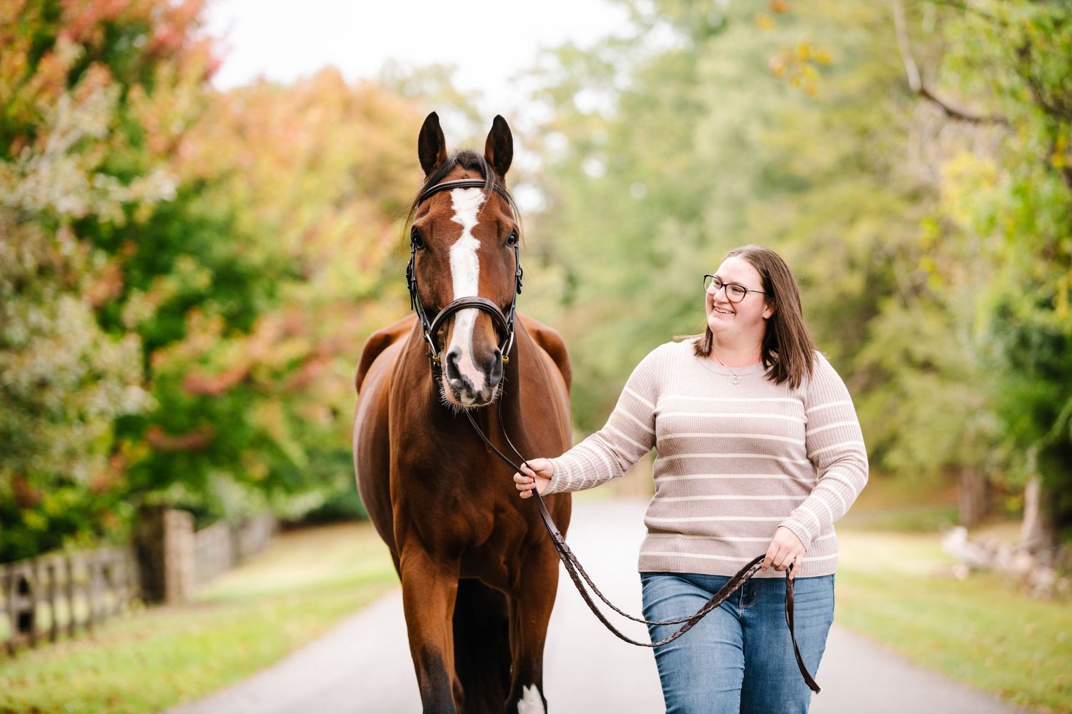 A bay horse with bridle being led by someone in a beige sweater on an autumn path.