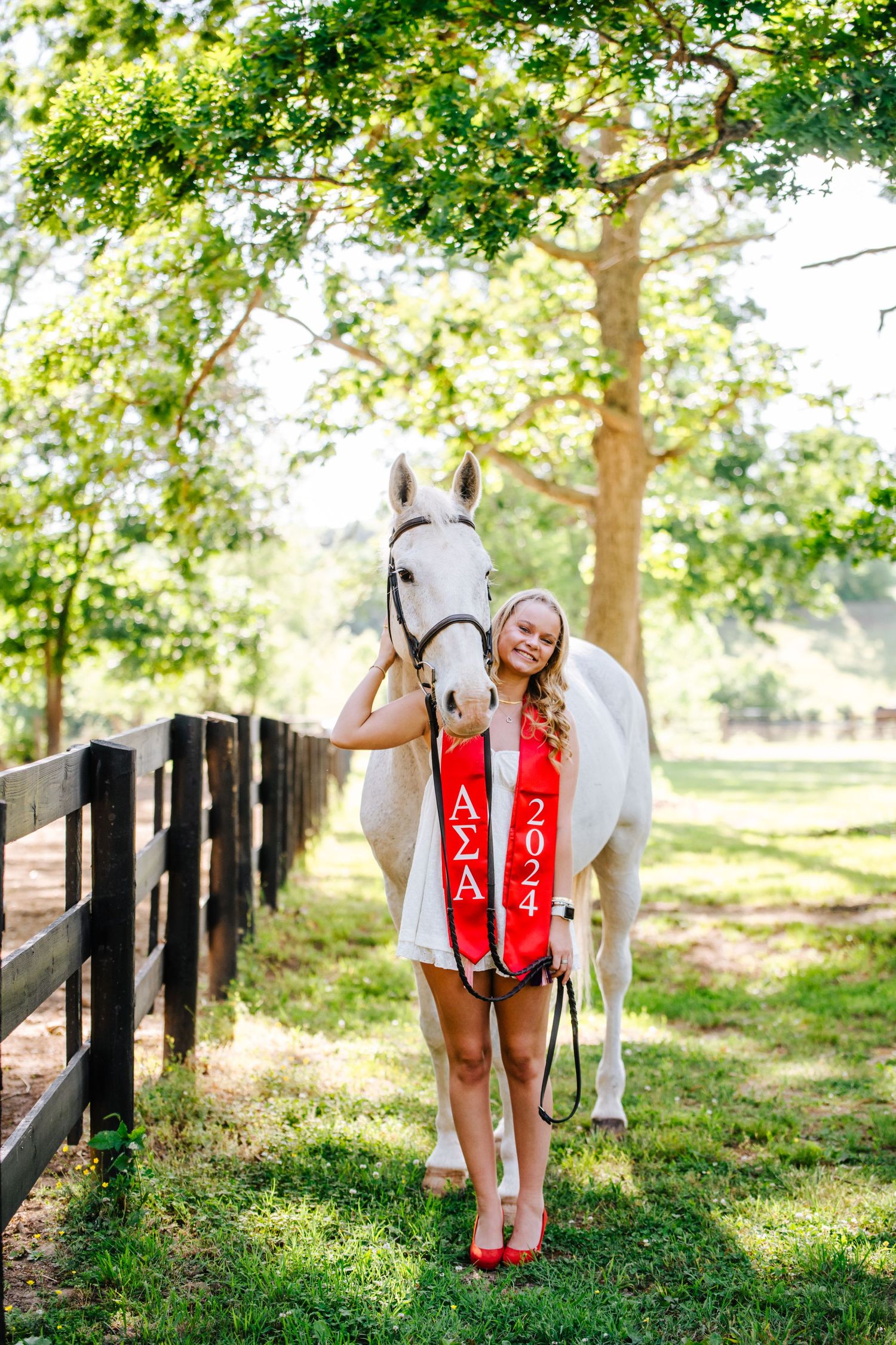 Student athlete poses with a white horse by a wooden fence in a sunny outdoor setting wearing formal attire and sash.