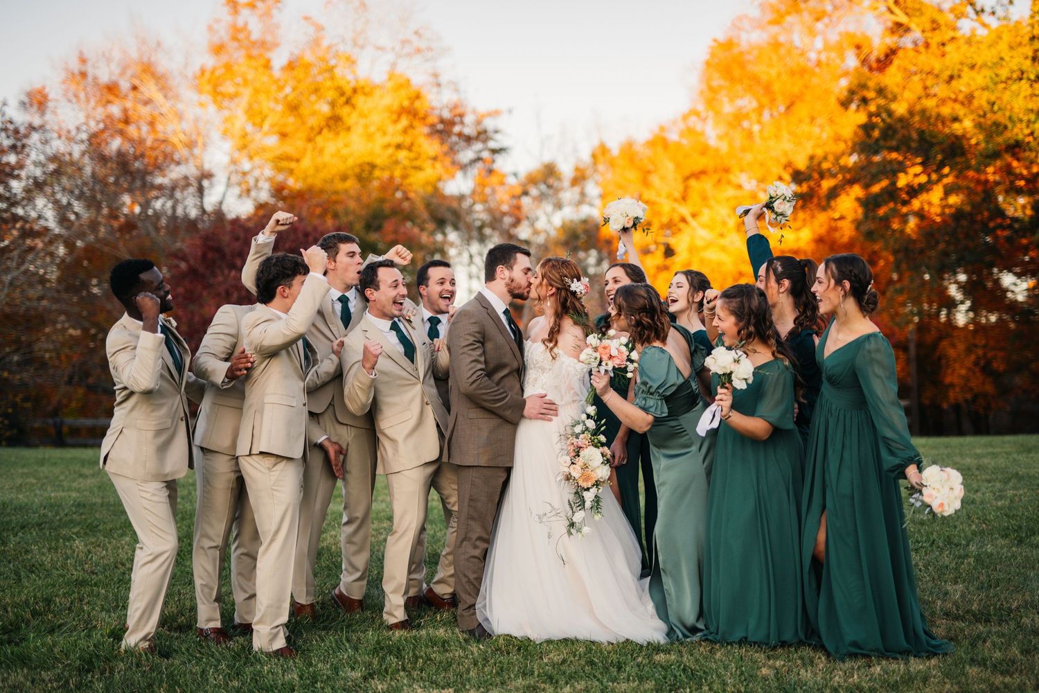 Wedding party in green dresses and beige suits posed together against vibrant autumn foliage.