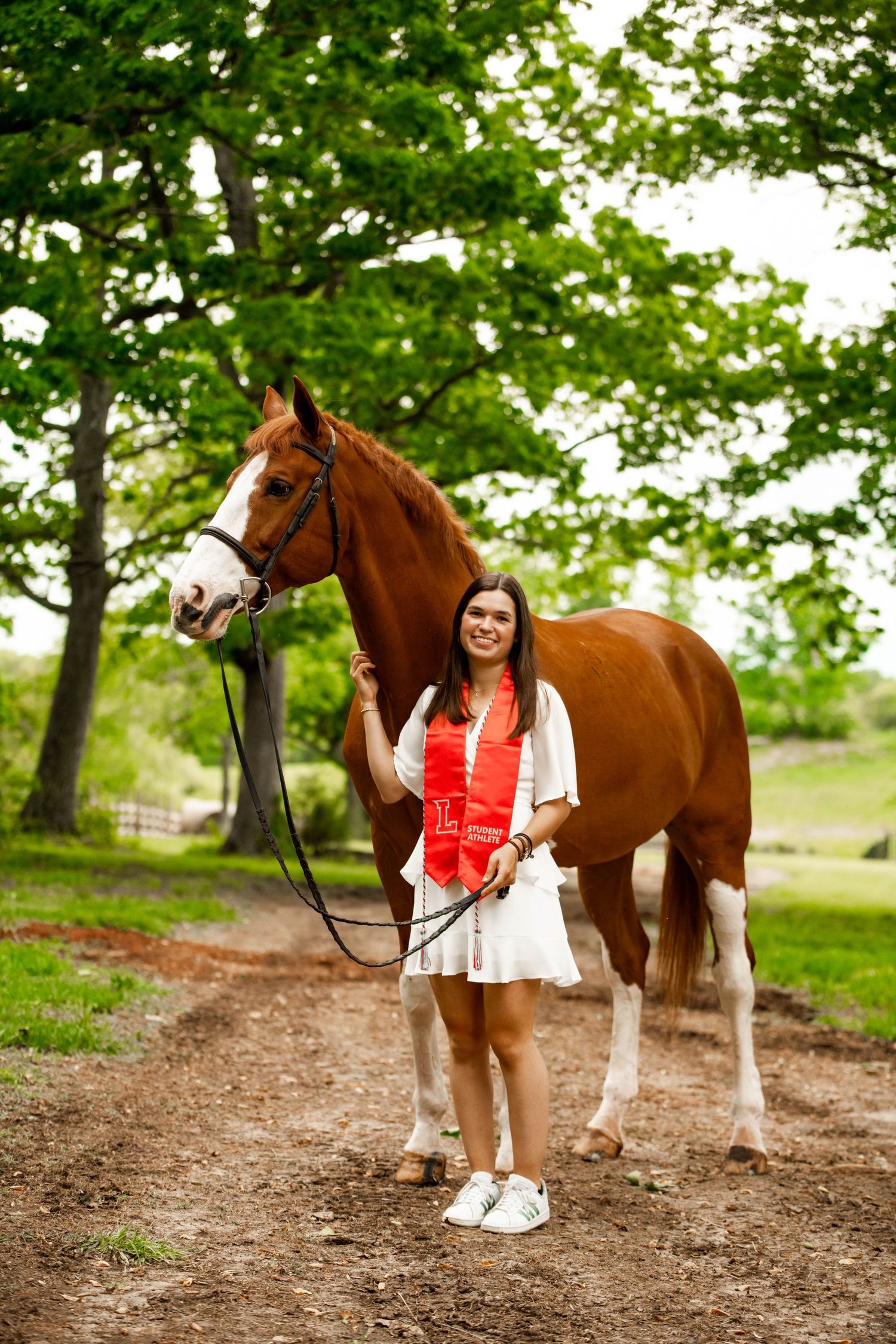 A chestnut and white horse stands with graduate on a dirt path surrounded by trees.