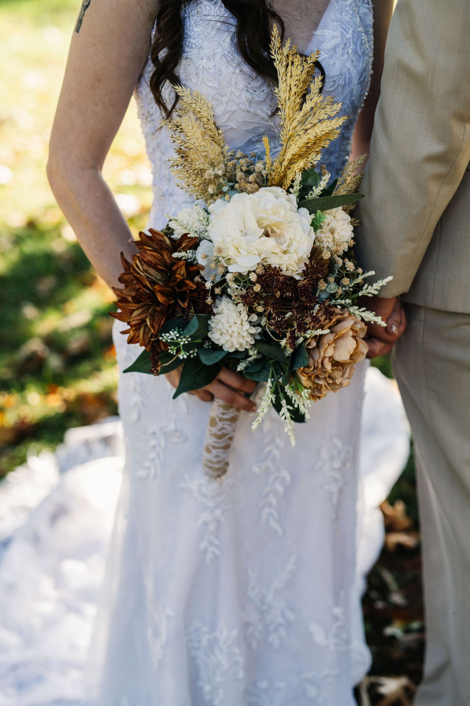 A close-up of a bridal bouquet featuring white flowers and golden accents against a lace dress.