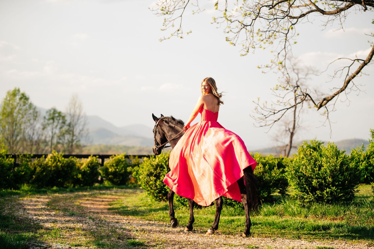 A rider in a flowing red dress sits atop a dark horse on a country path during springtime.