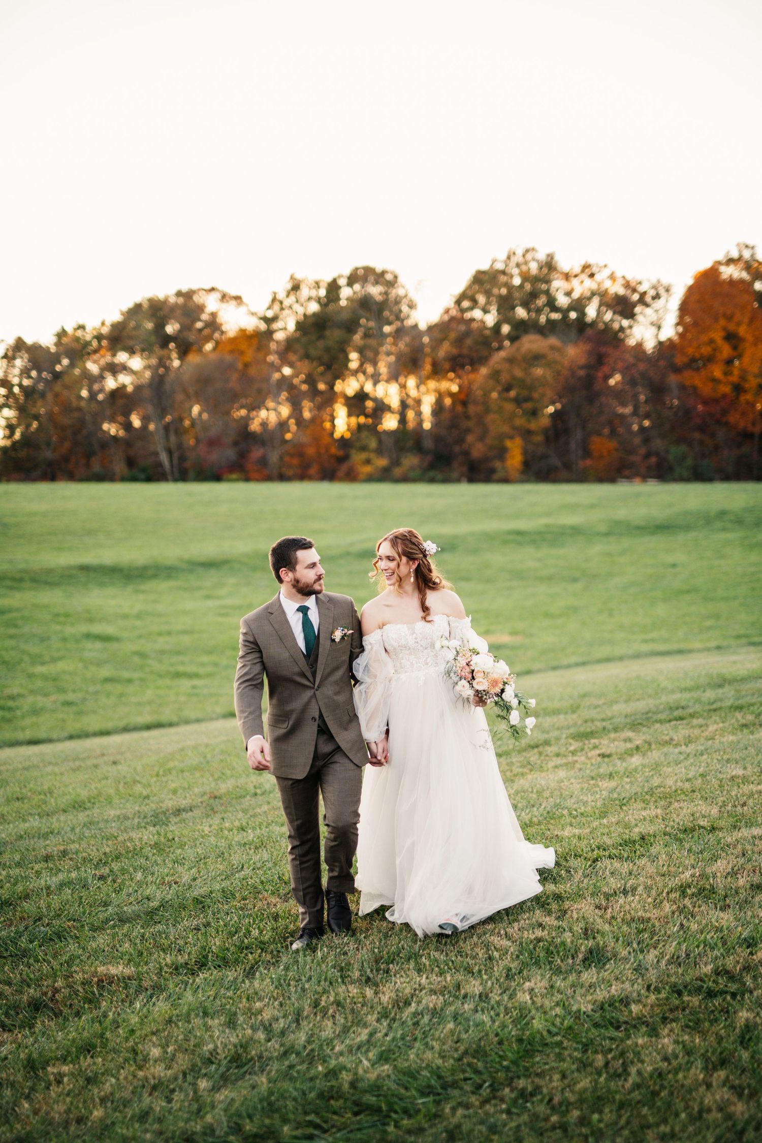 Newlyweds walk hand in hand across a hillside during sunset.