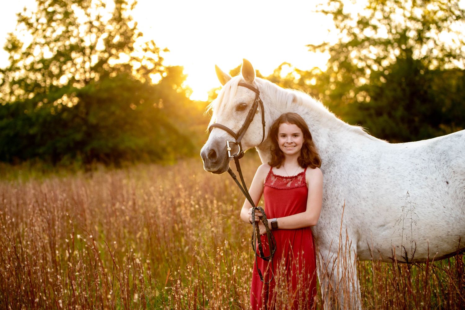 A white horse and companion in a red dress stand in a field during golden hour sunset.