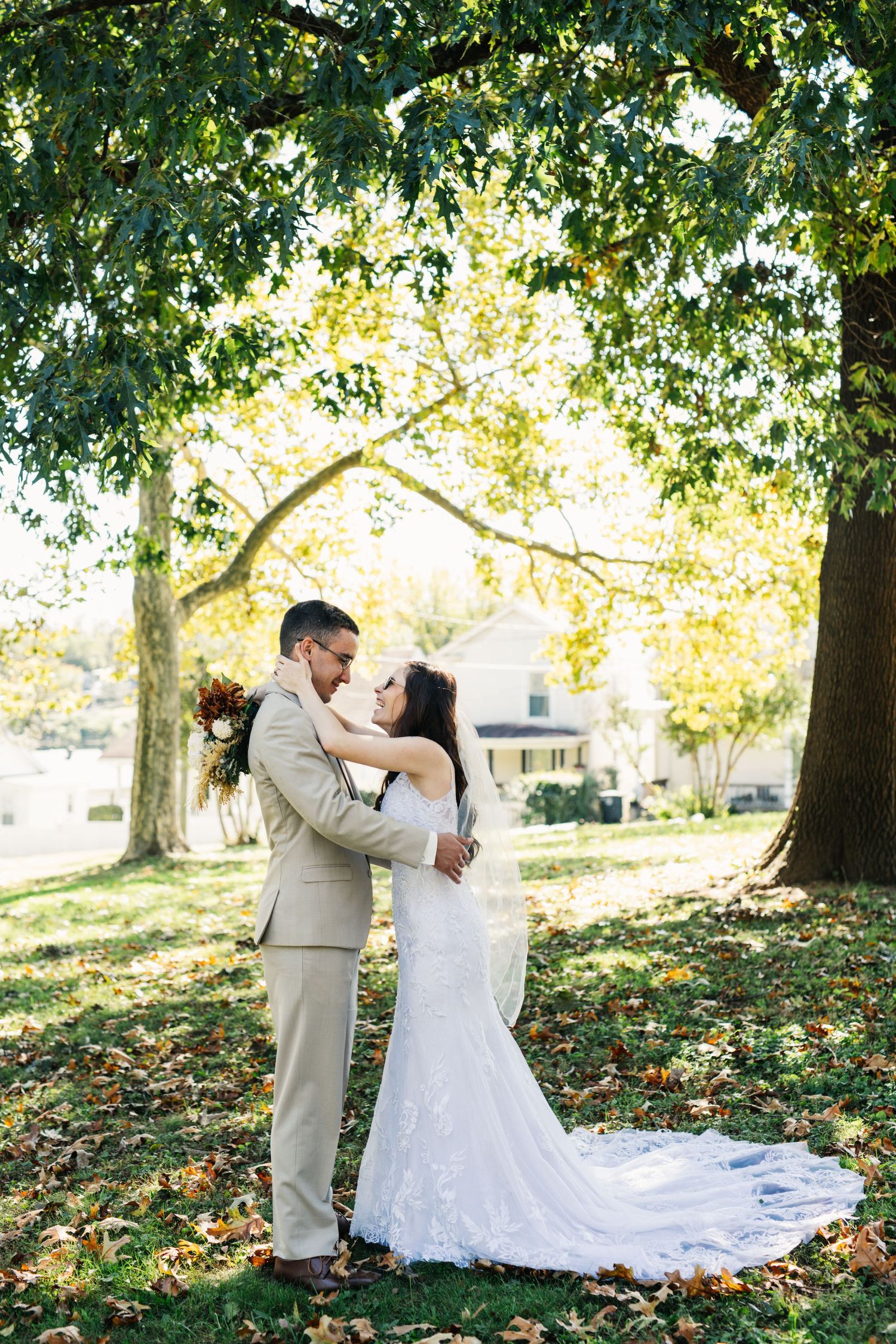 A wedding couple embraces on fallen autumn leaves under large trees.