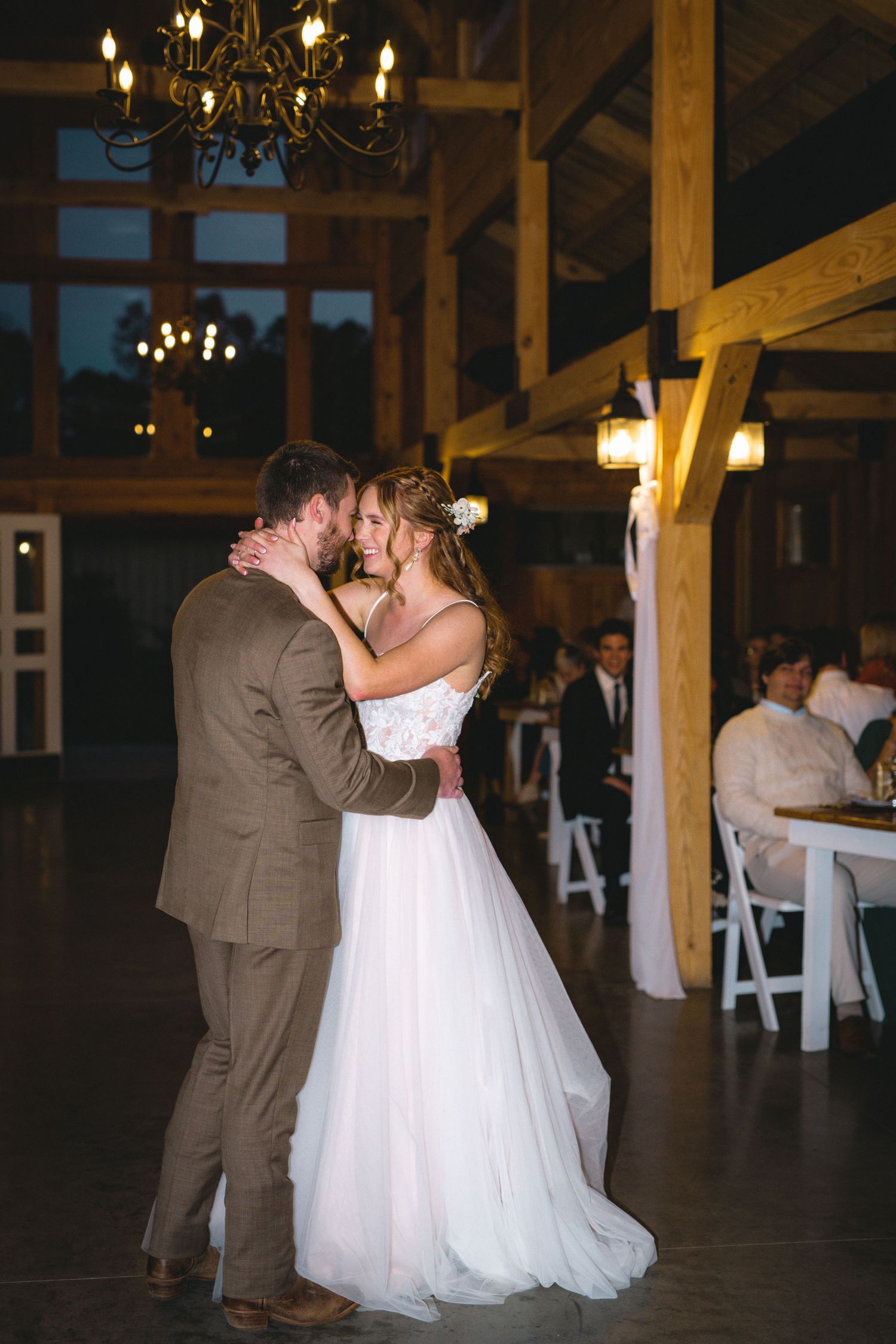 A romantic first dance in a rustic barn venue with wooden beams and chandeliers overhead.